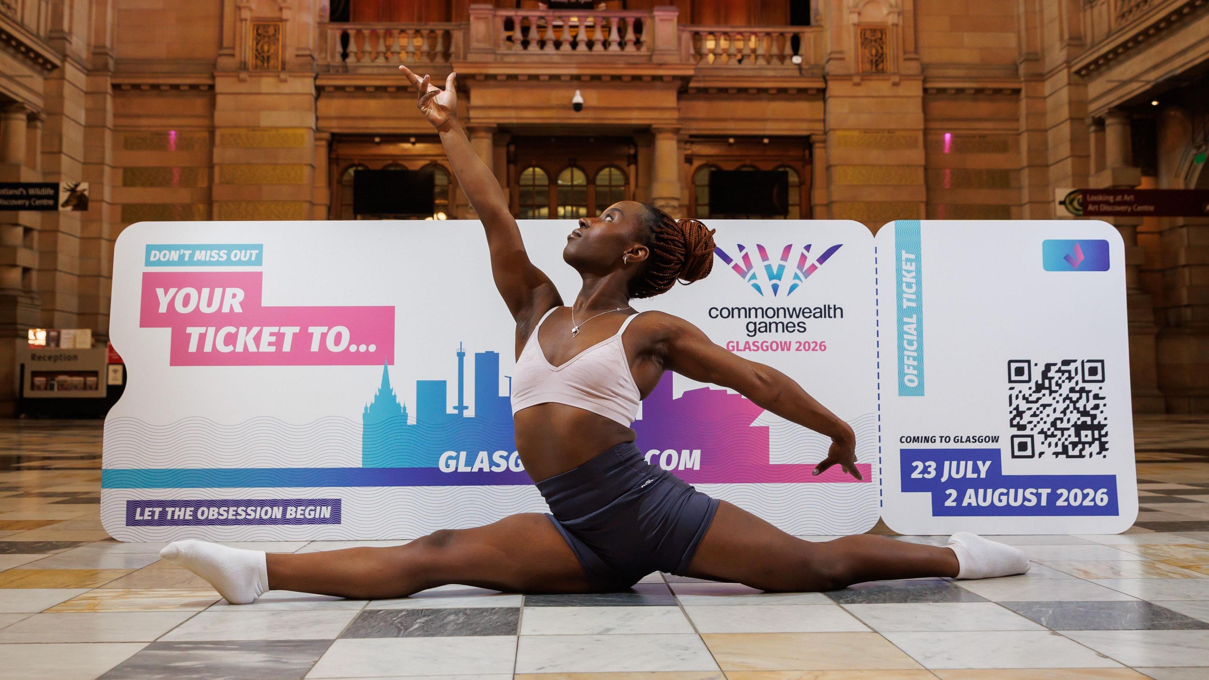 Gymnastics fan Mercy Abel, a young woman in shorts and a crop top, sits in splits in front of a giant Glasgow 2026 ticket mock-up. The photo is taken in the grand hall at Kelvingrove Art Gallery.
