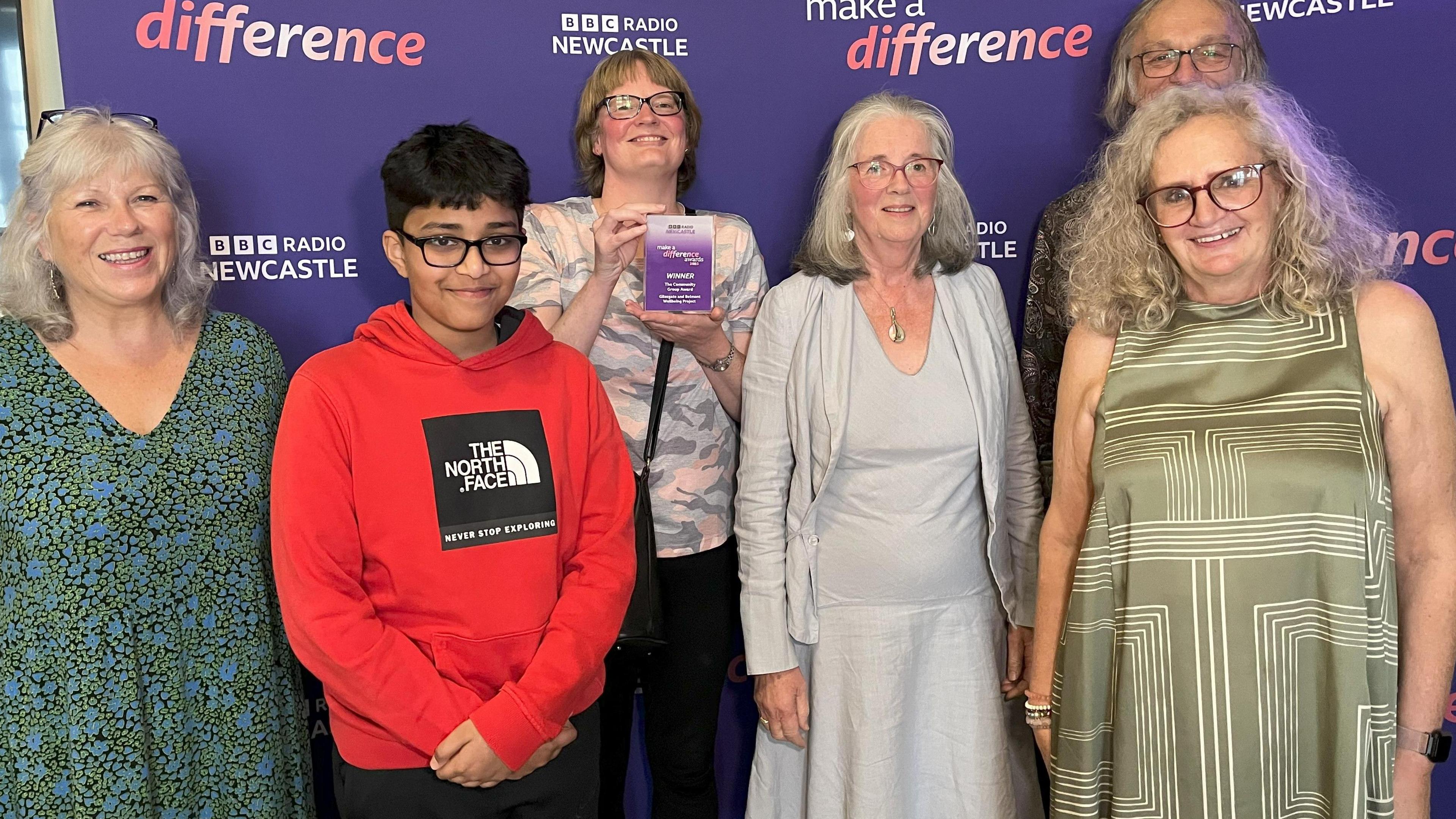 Volunteers from the Gilesgate and Belmont Wellbeing Project. They are standing in front of purple signage that reads 'MAKE A DIFFERENCE' with the BBC Radio Newcastle branding. There are six volunteers of different ages and ethnicities.