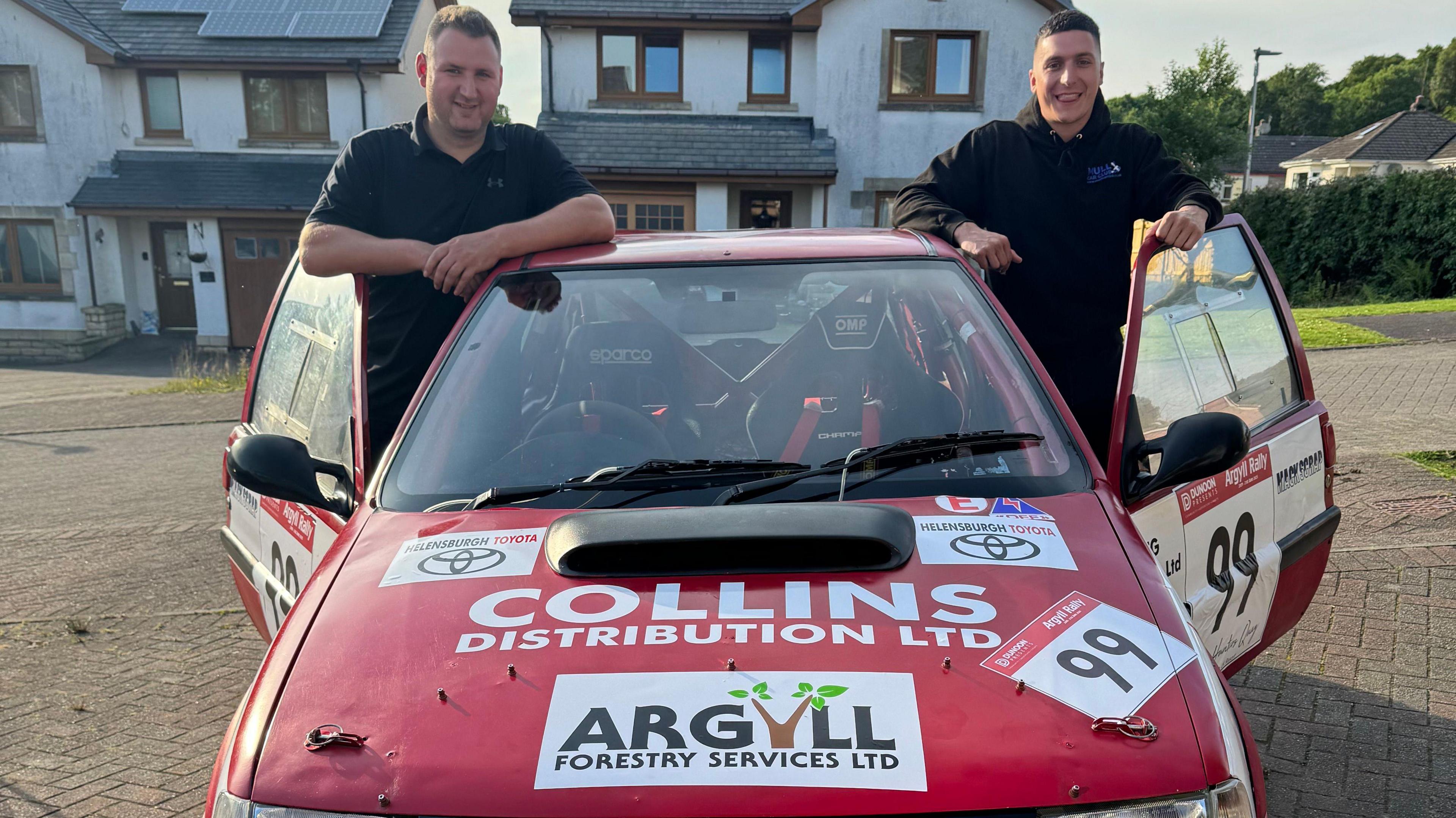 Two men, leaning on the open doors of a red car. It has sponsorship stickers on the bonnet. Both men are wearing black and smiling.