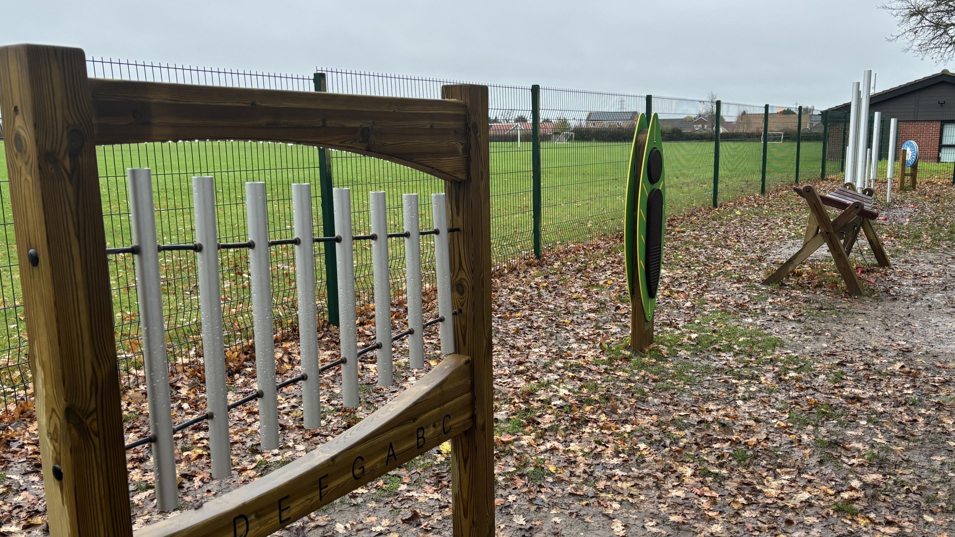 The image shows an outdoor musical play area set along a fenced boundary of a grassy field.