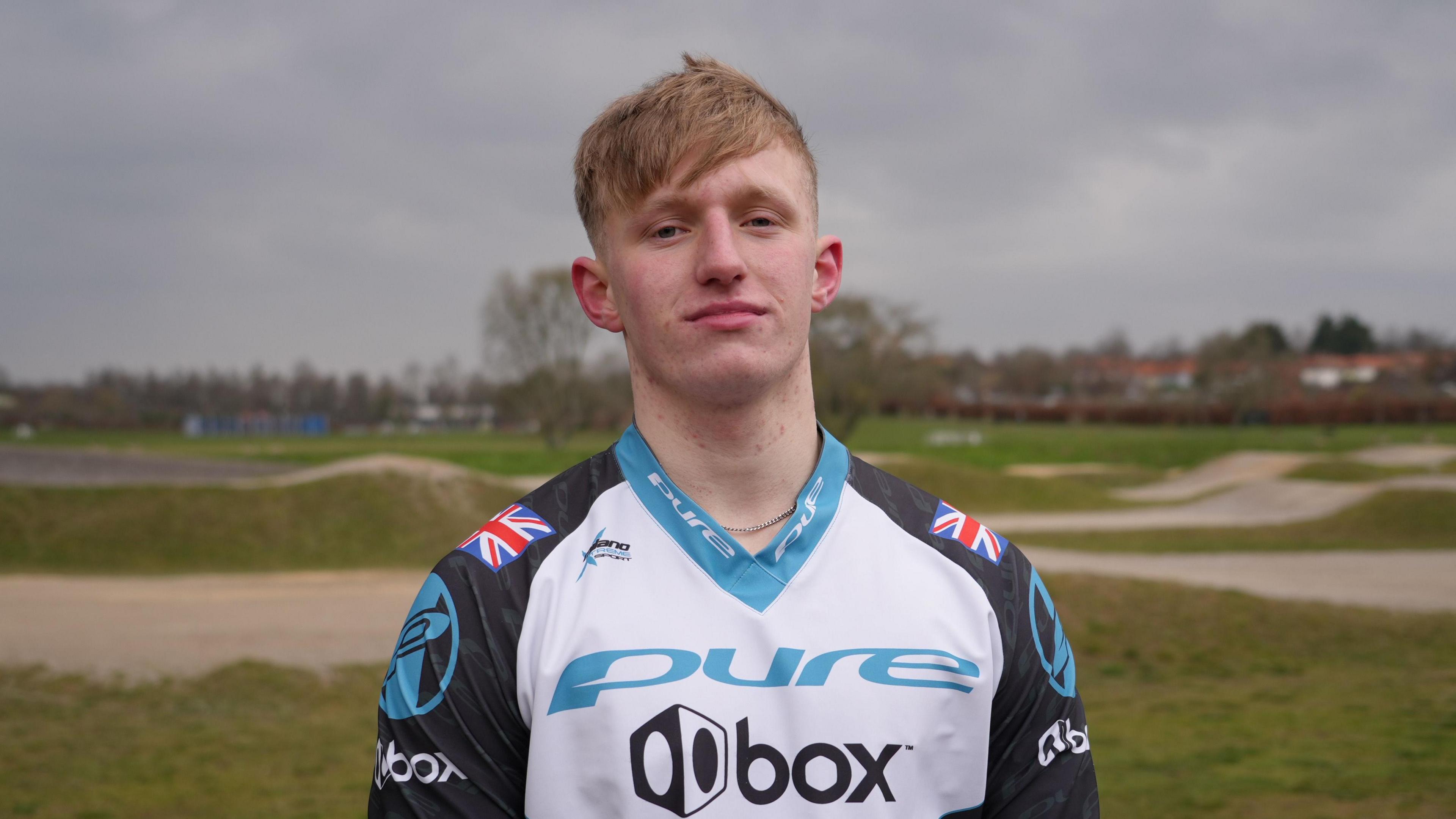 Jared Hill stands in front of a BMX track. He is wearing a V-necked, white, black and sky blue racing jersey with Union flags on each shoulder and the word "Pure" across the chest.