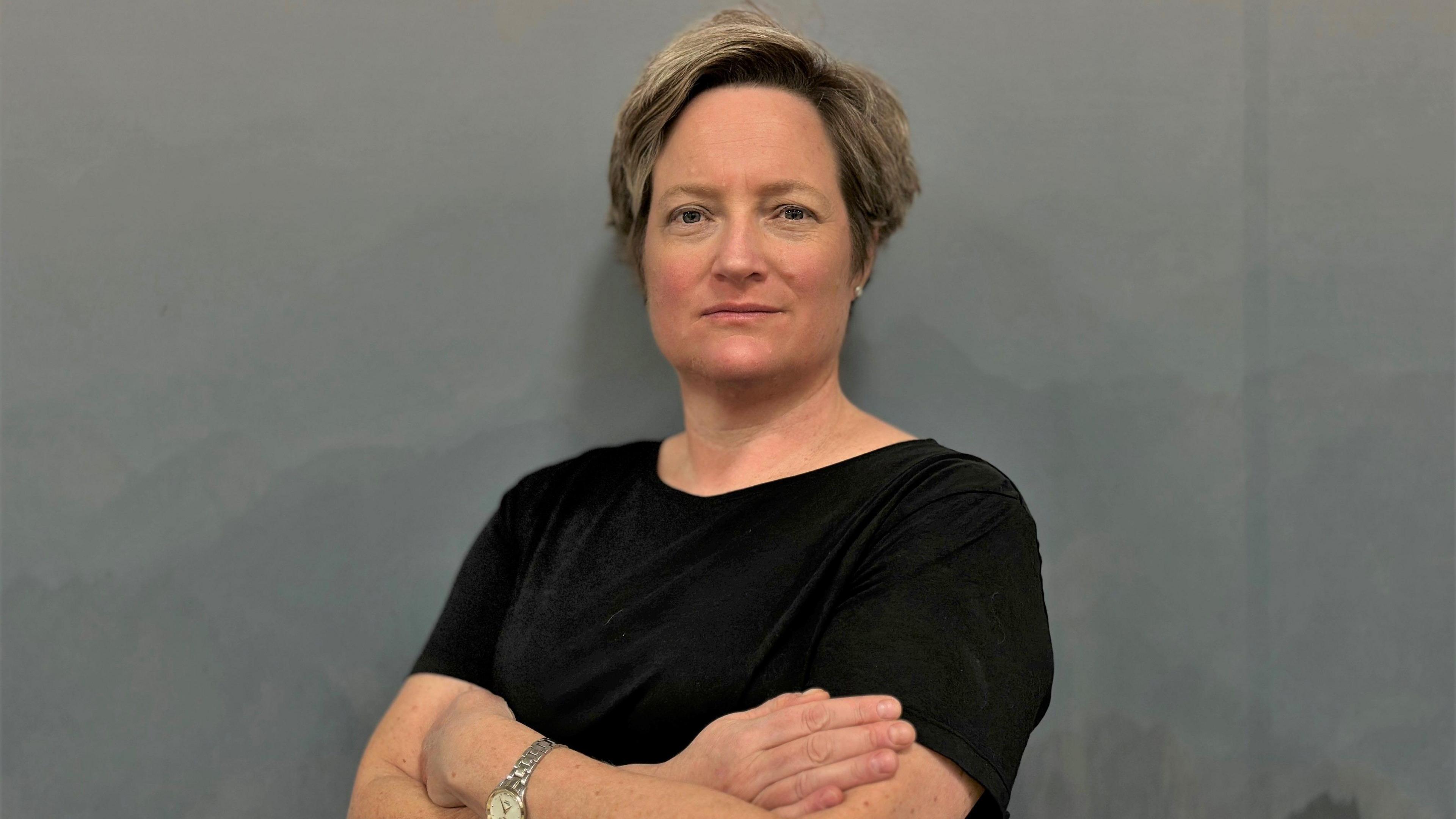 Claire Stuckey standing against a dark grey background with her arms folded. She is looking at the camera with a determined expression. She is wearing a black t-shirt, a silver watch, and has short brown hair in a side parting.