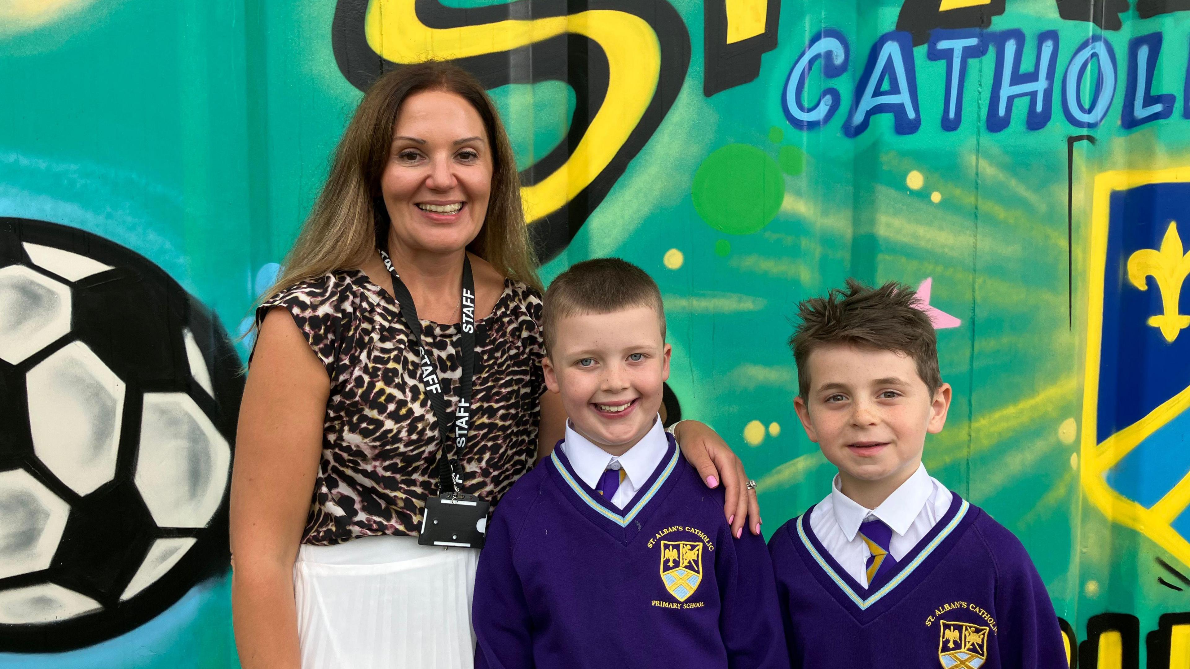 A woman with brown hair, wearing a leopard print top and white skirt, stands next to two boys in school uniform. They all smile at the camera and stand in front of a school mural.