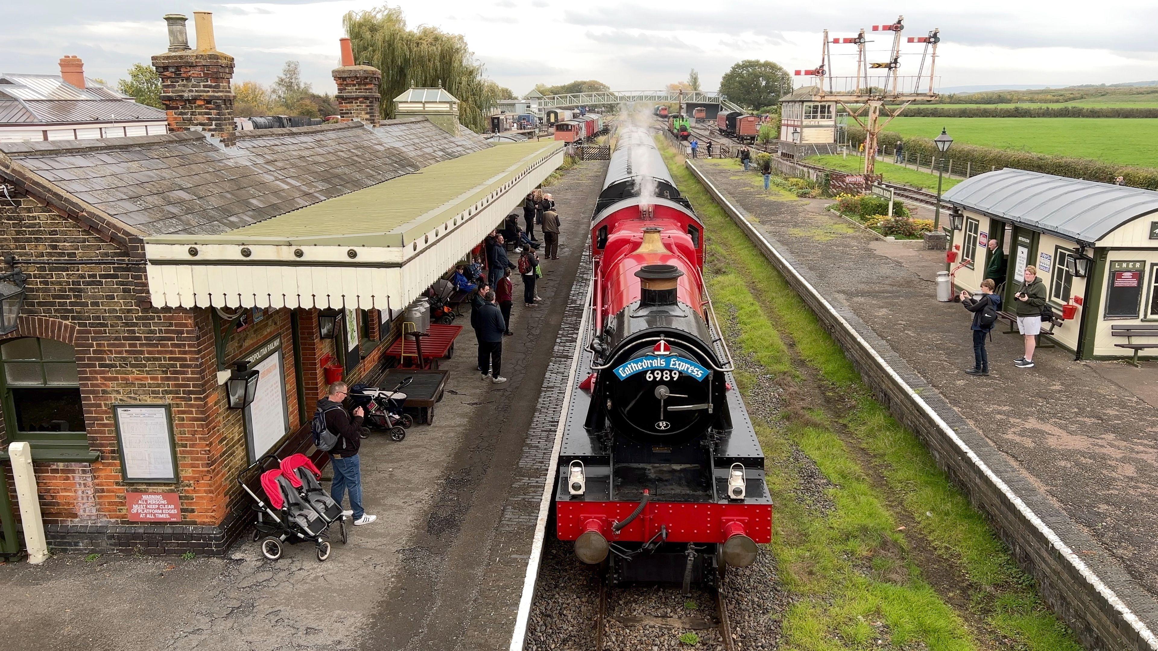 An aerial view, or likely taken from a railway bridge. It shows the steam engine arriving at the railway station, with steam billowing, and people on the platforms taking photos. It is a cloudy day.