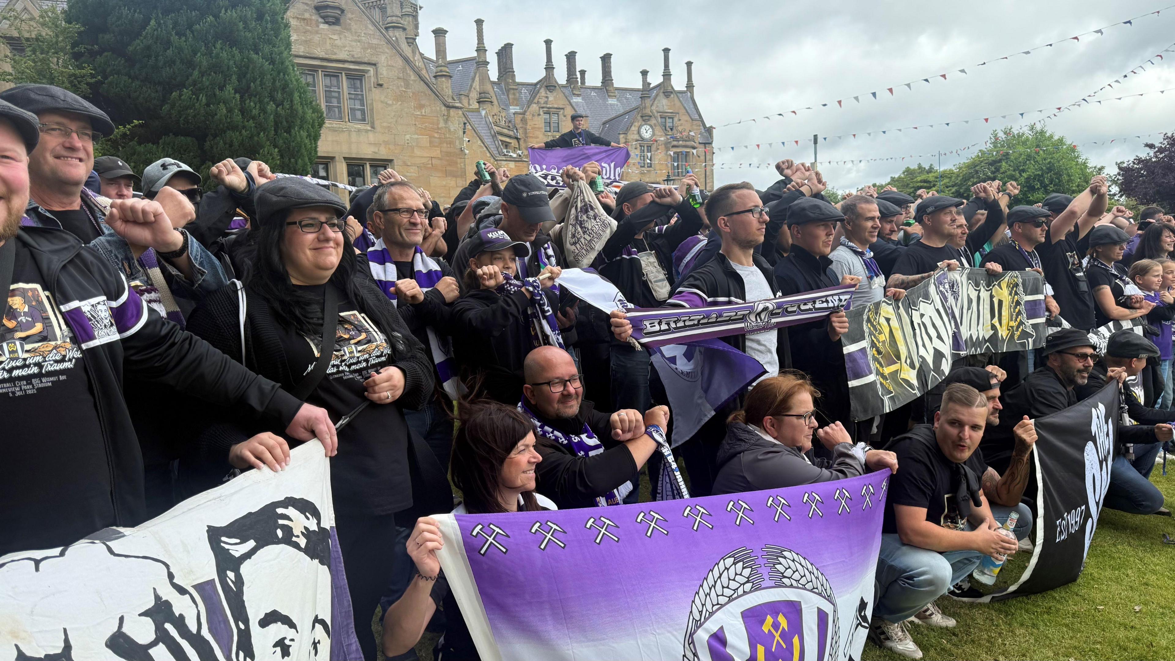 A large group of Erzgebirge Aue fans holding flags and banners standing outside Brownlow House - a brown brick building in a Baronial style