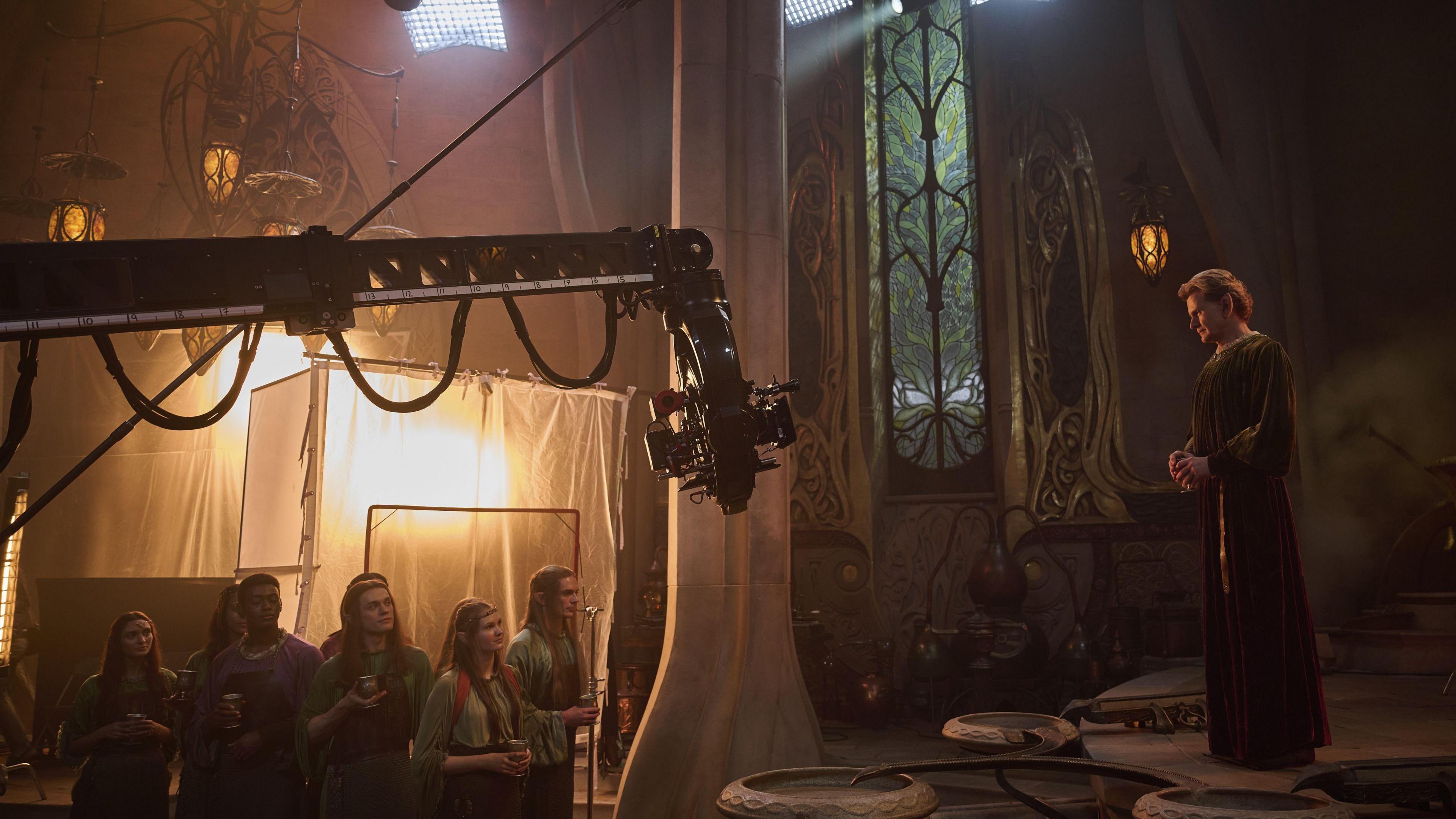 A behind the scenes shot of character Celebrimbor from Rings of Power in his forge. There are high ceilings, warm lighting and a group of elves on a platform lower than him