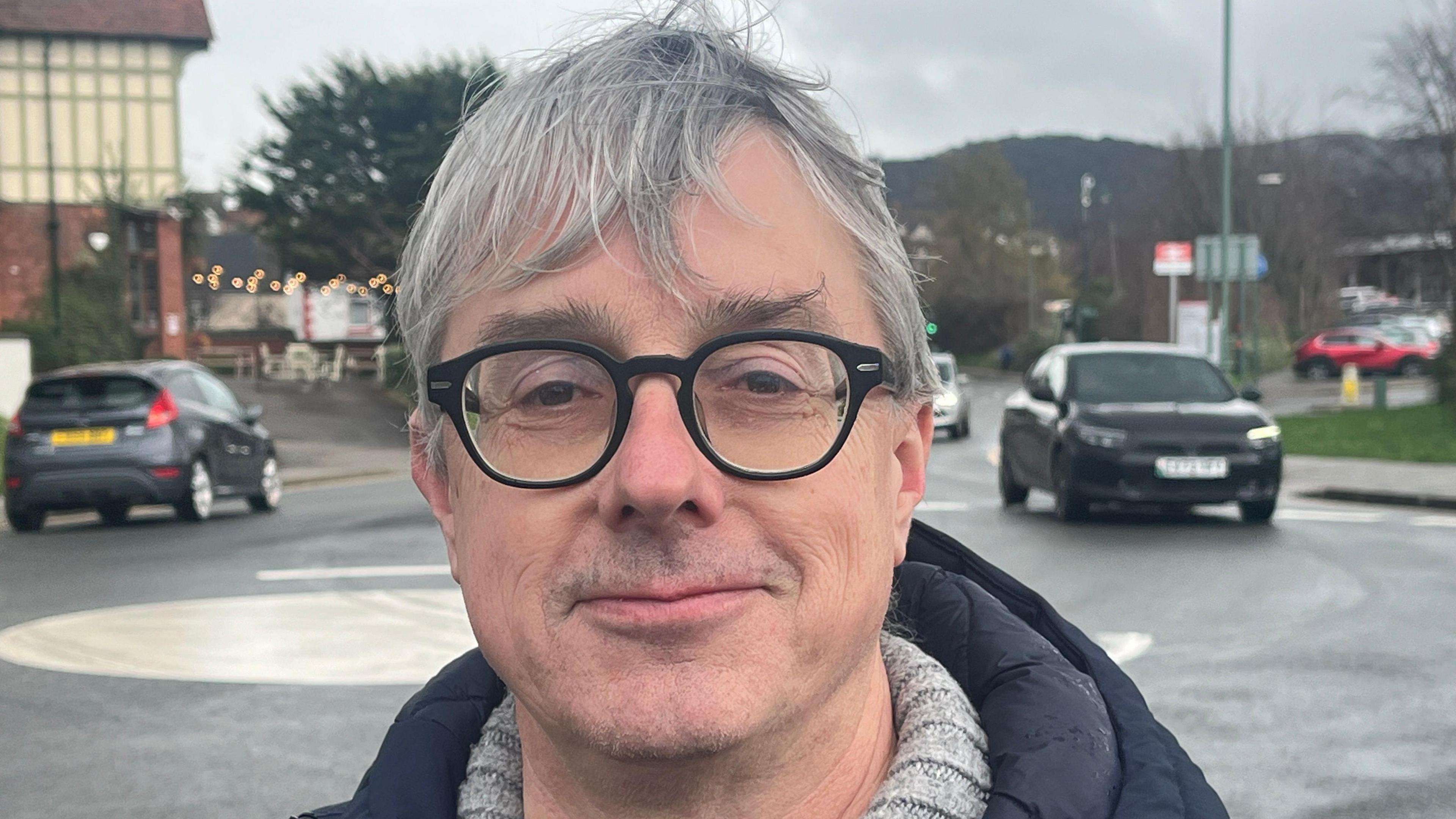 Rhodri Clarke mewn sbectol a chot ddu