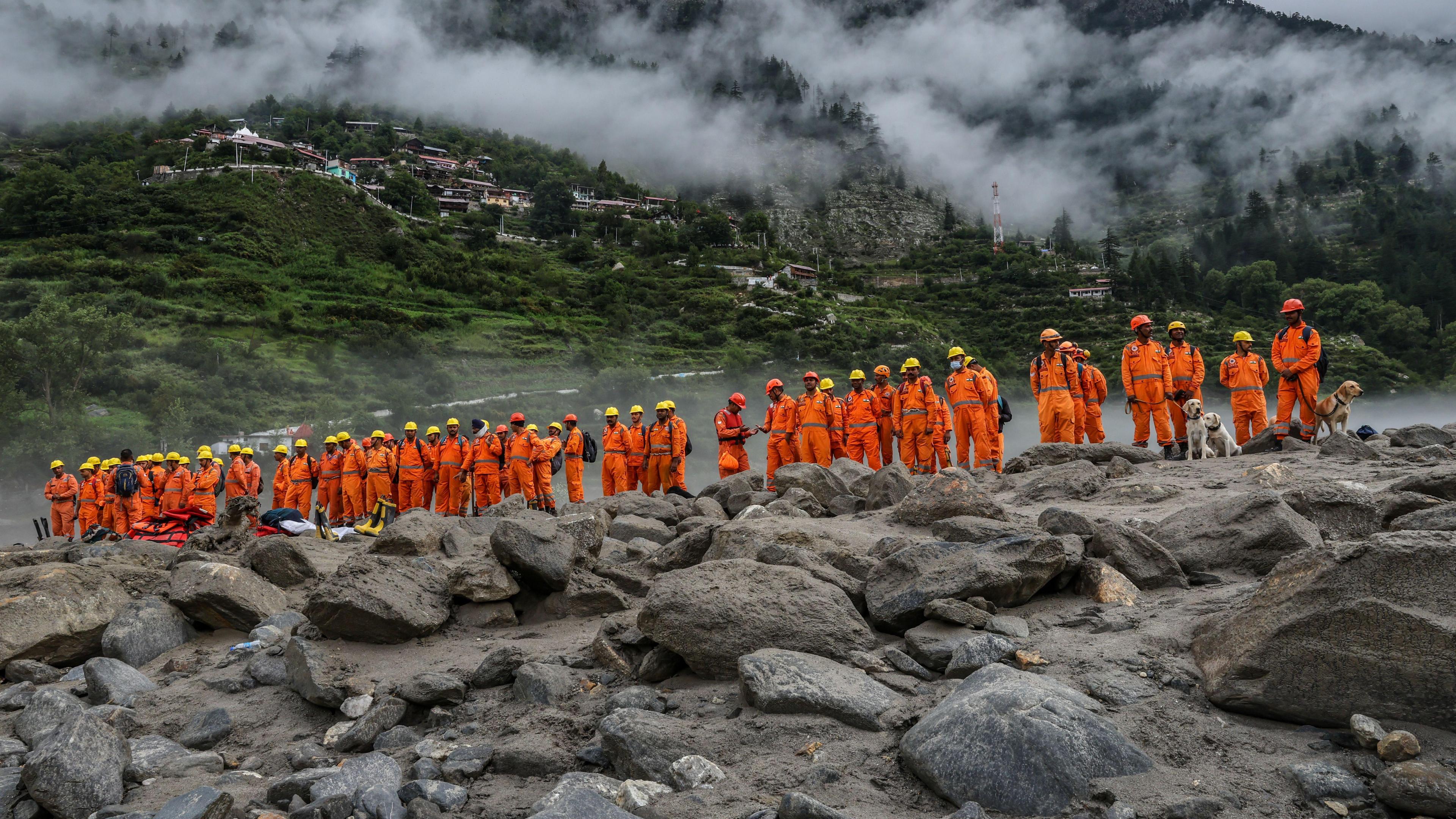 Heavy rainfall in several parts of the country has caused landslides and floods, inundating villages and towns and killing hundreds. A group of rescue workers in orange standing at a rocky terrain with sniffer dogs. A misty hillock with buildings can be seen behind them.