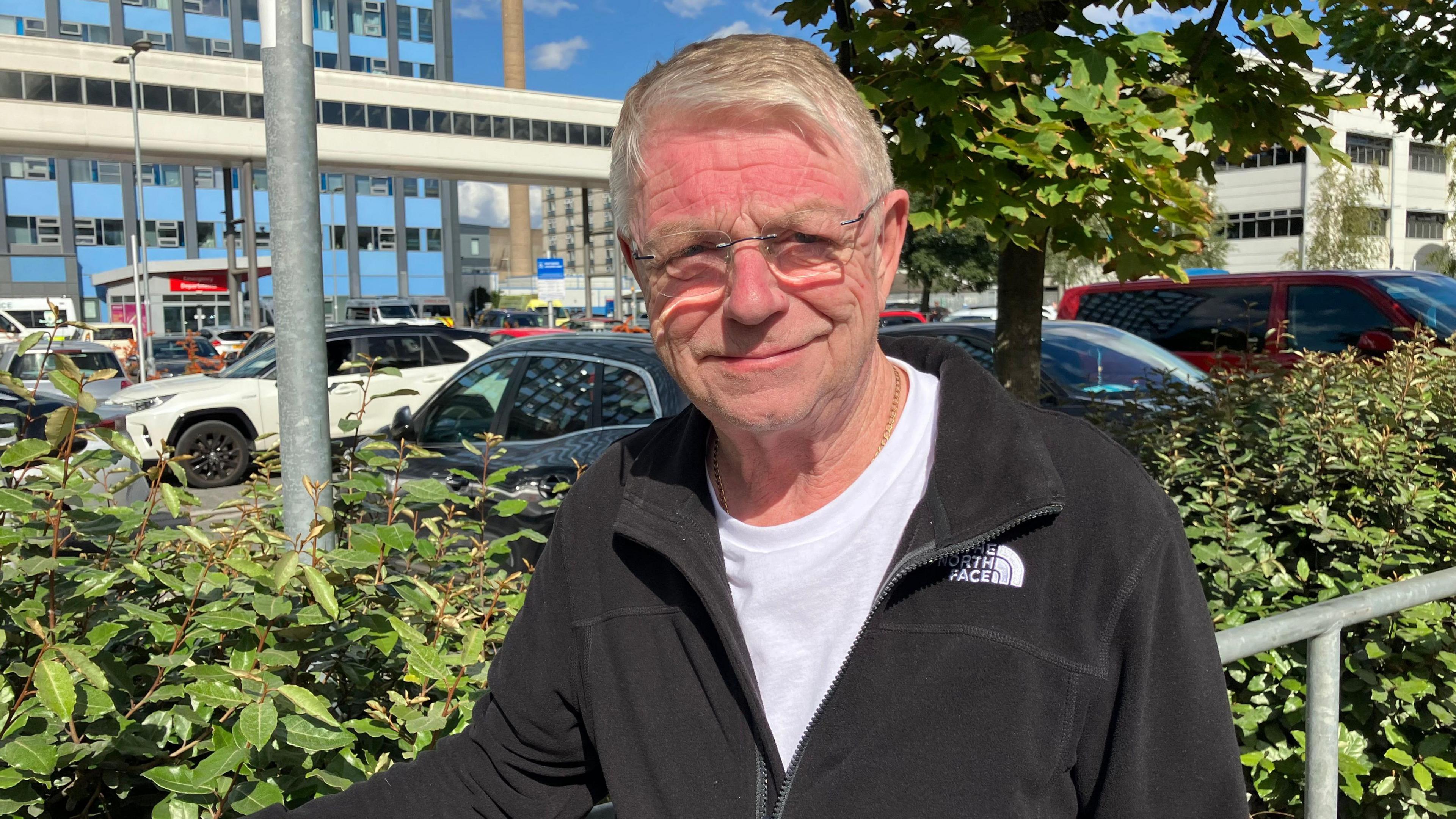 A man with grey hair and rectangular clear glasses is wearing a white top and black North Face fleece and looking at the camera smiling. He is leaning against a railing and you can see a car park behind him and a building with several floors.