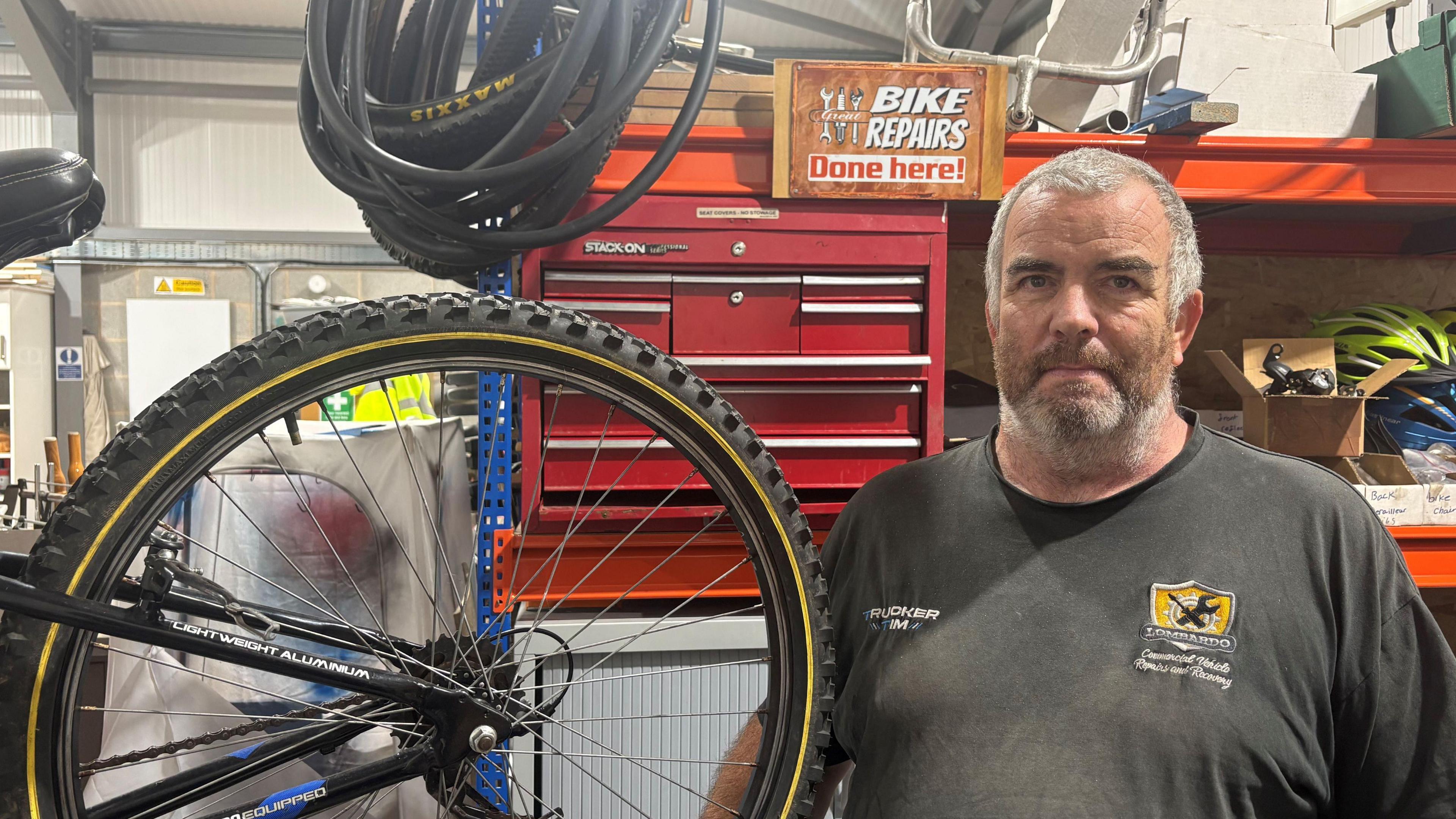 Craig Brown, also known as Buster, stood next to a black bike in front of a work unit with tools and equipment on. He has short grey hair and grey facial hair. He is wearing a black t-shirt with a yellow logo on.