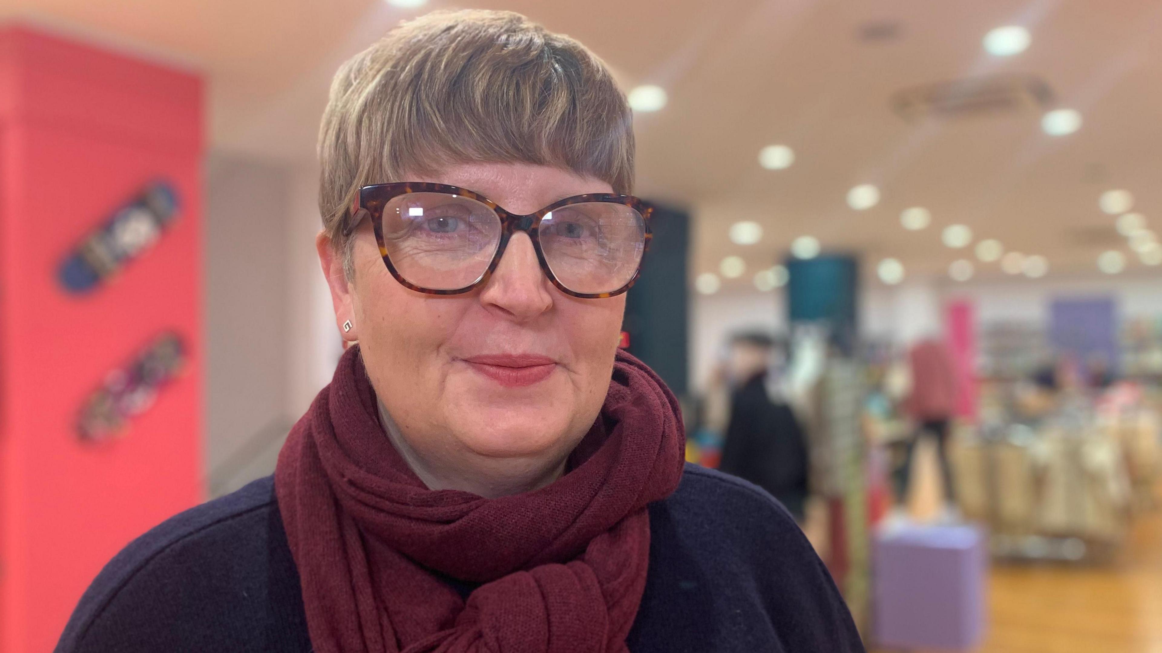 SJ Waites looking at the camera wearing glasses, a burgundy scarf and a navy top.