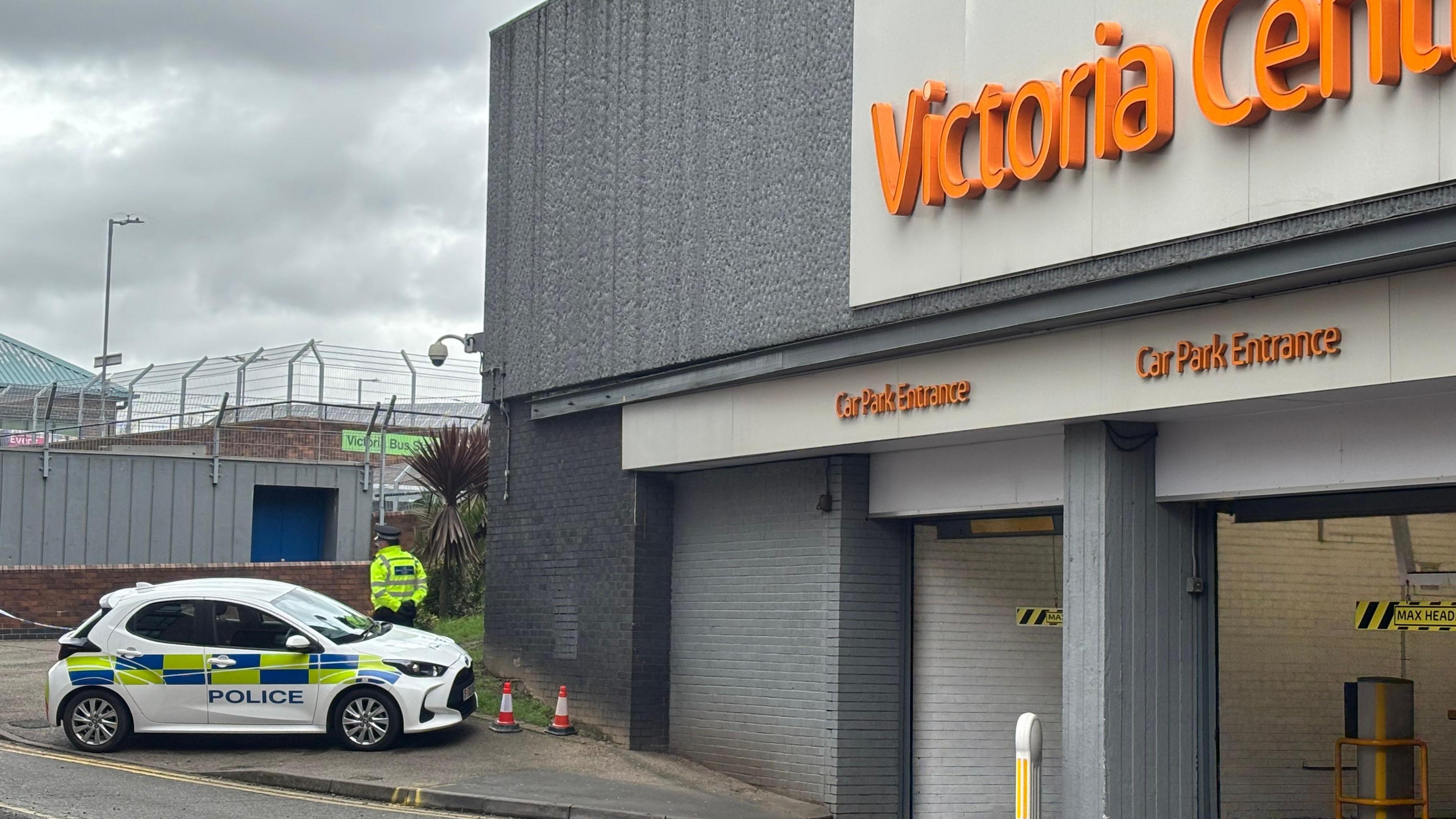 A small police vehicle is parked on the pavement next to the entrance to a large grey car park building. Some police tape is to the left of the car and a police officer in hi-vis clothing is stood facing away from the car. The bus station building is in the background.