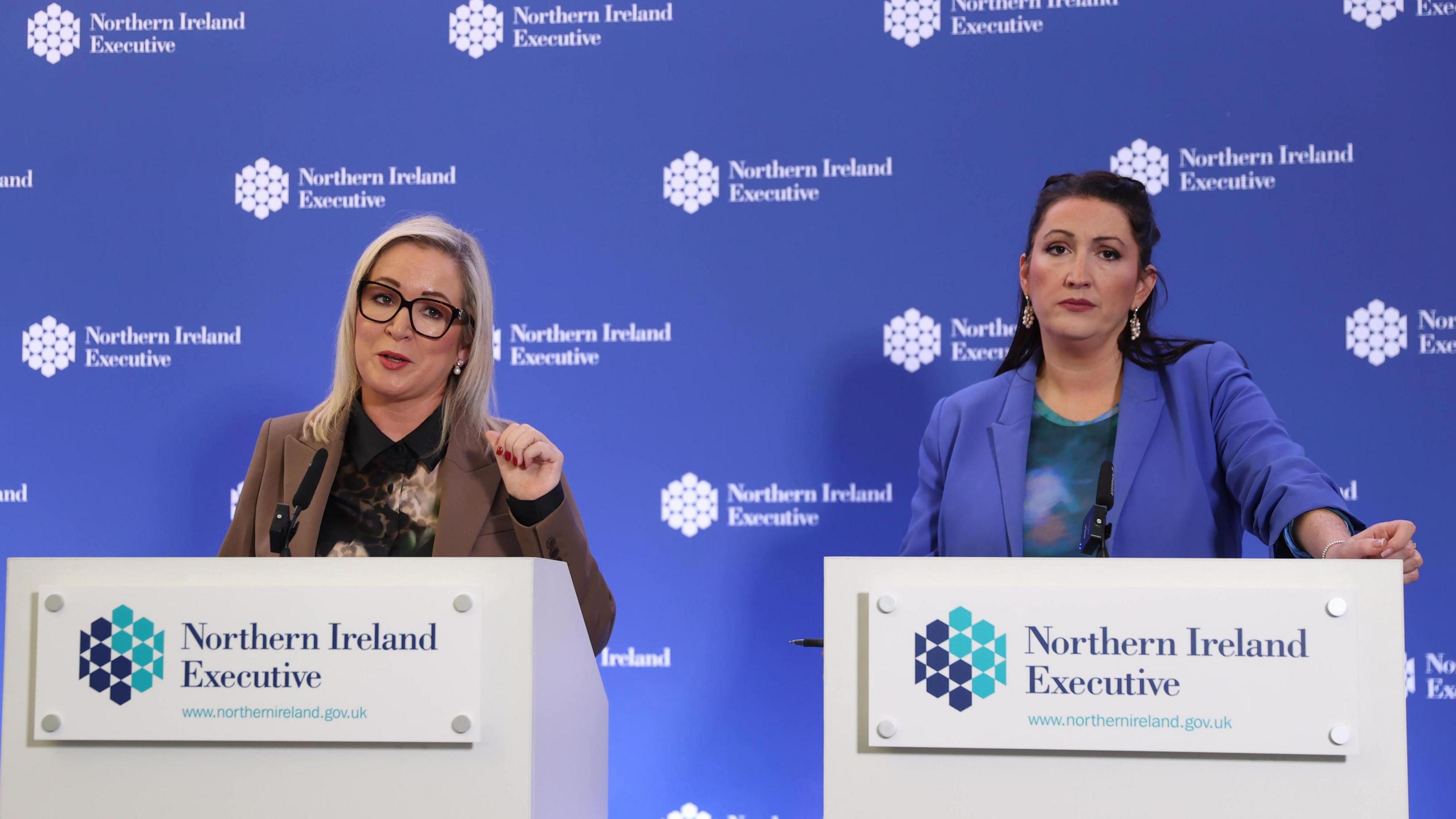 Two women stand at lecterns in front of a blue background. The blue background has the words 'Northern Ireland Executive' written on it multiple times. The same words are written on both lecterns. The woman on the left has blonde hair and is wearing glasses. She is wearing a dark shirt and a brown blazer. The woman on the right is wearing a green top and a lilac blazer. She has dark hair.