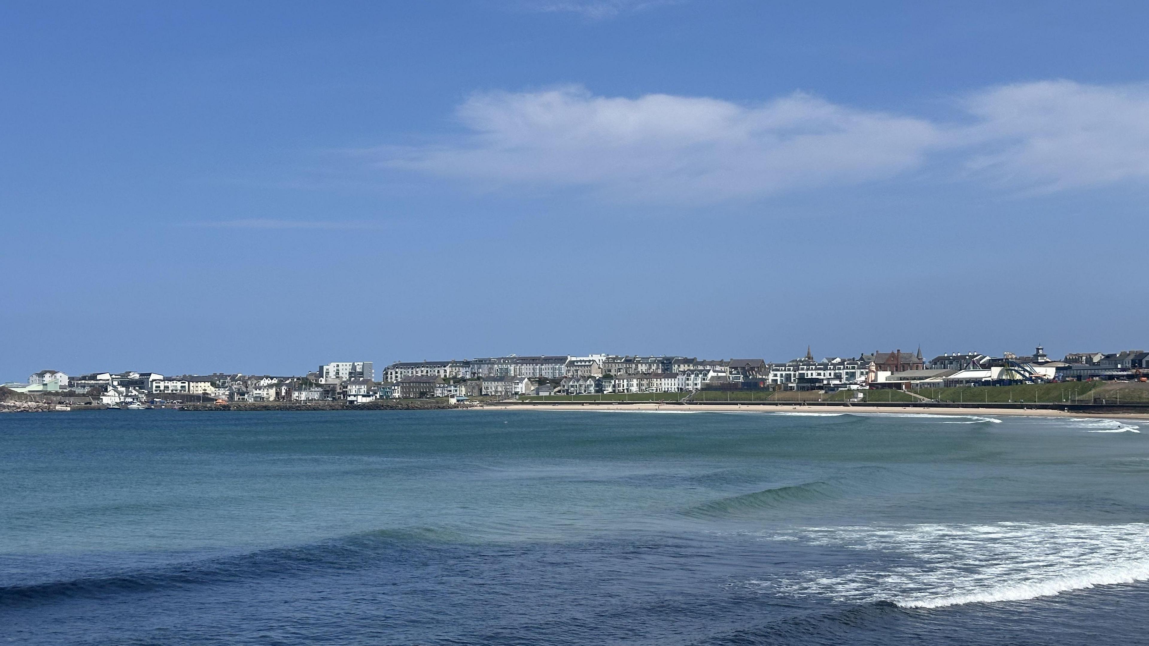 A scenic view overlooking the west strand in Portrush with apartments and homes far off in the distant. It is a sunny day with a blue sky and a blue sea with waves crashing.