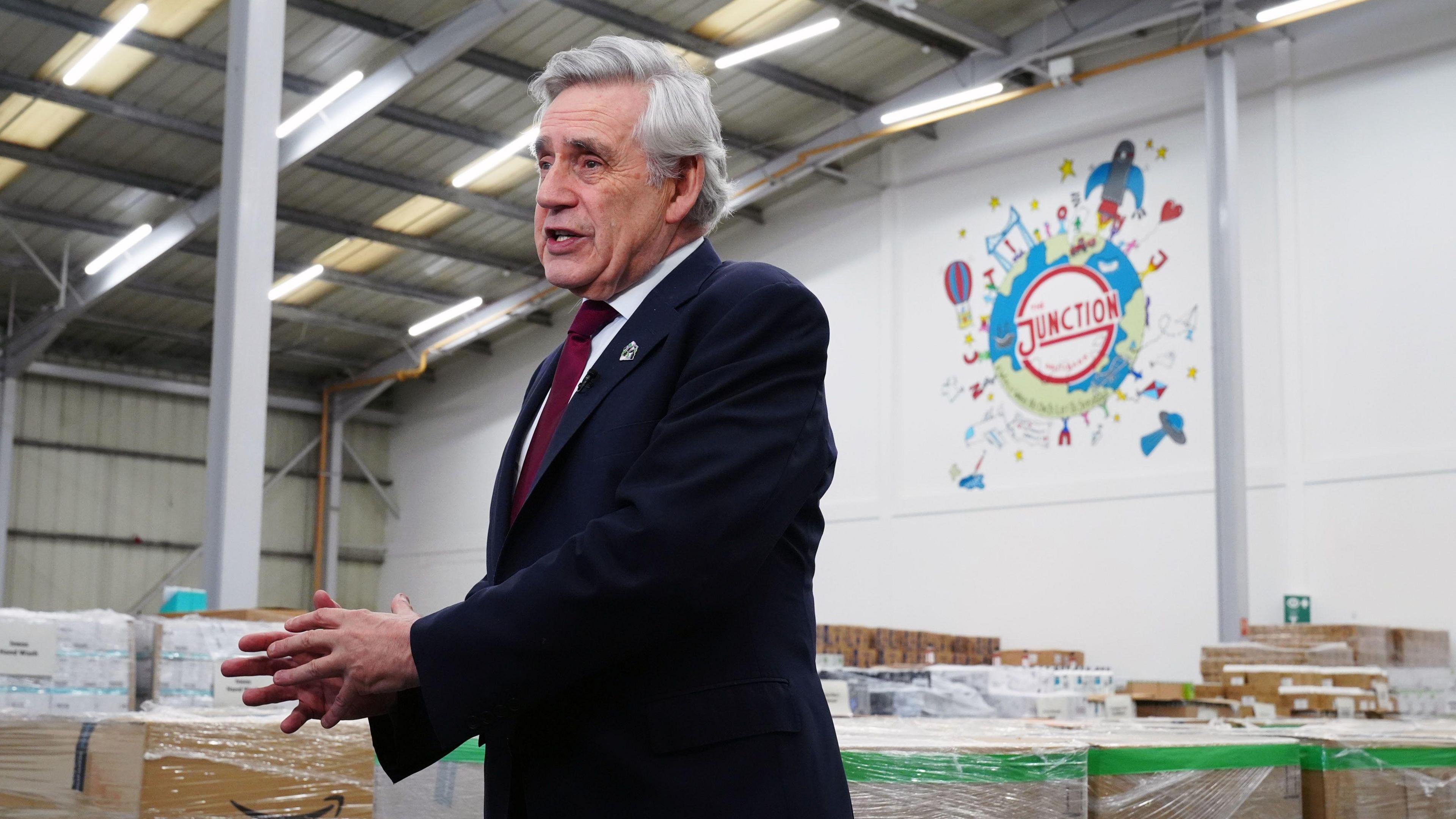 A man with grey hair and who is wearing a black suit jacket with a white shirt and red tie, in front of a warehouse full of boxes. 