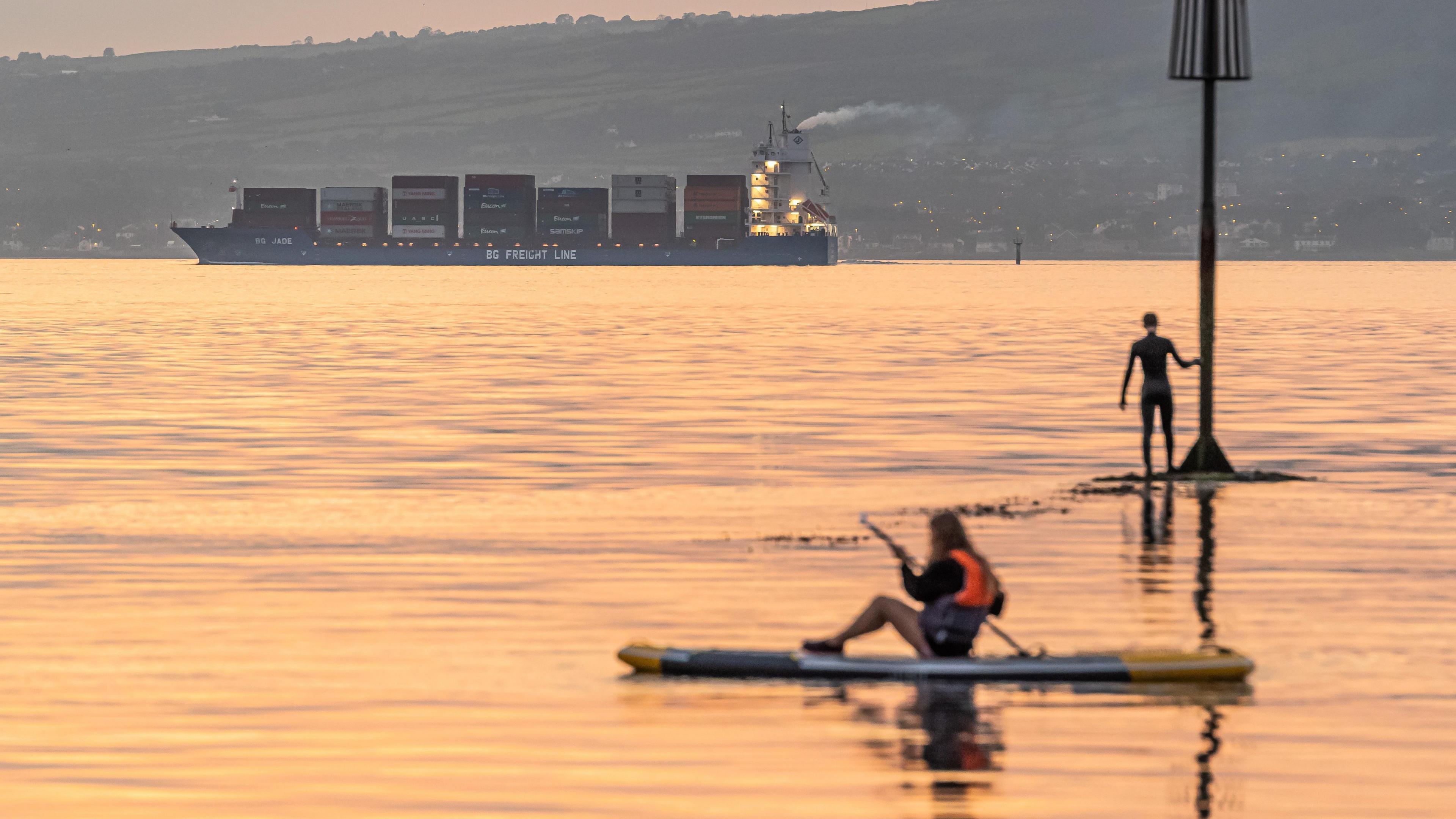 A small container ship sailing along Belfast Lough towards port as the sun sets across the lough. A woman is on a paddleboard and a man is standing in the water.