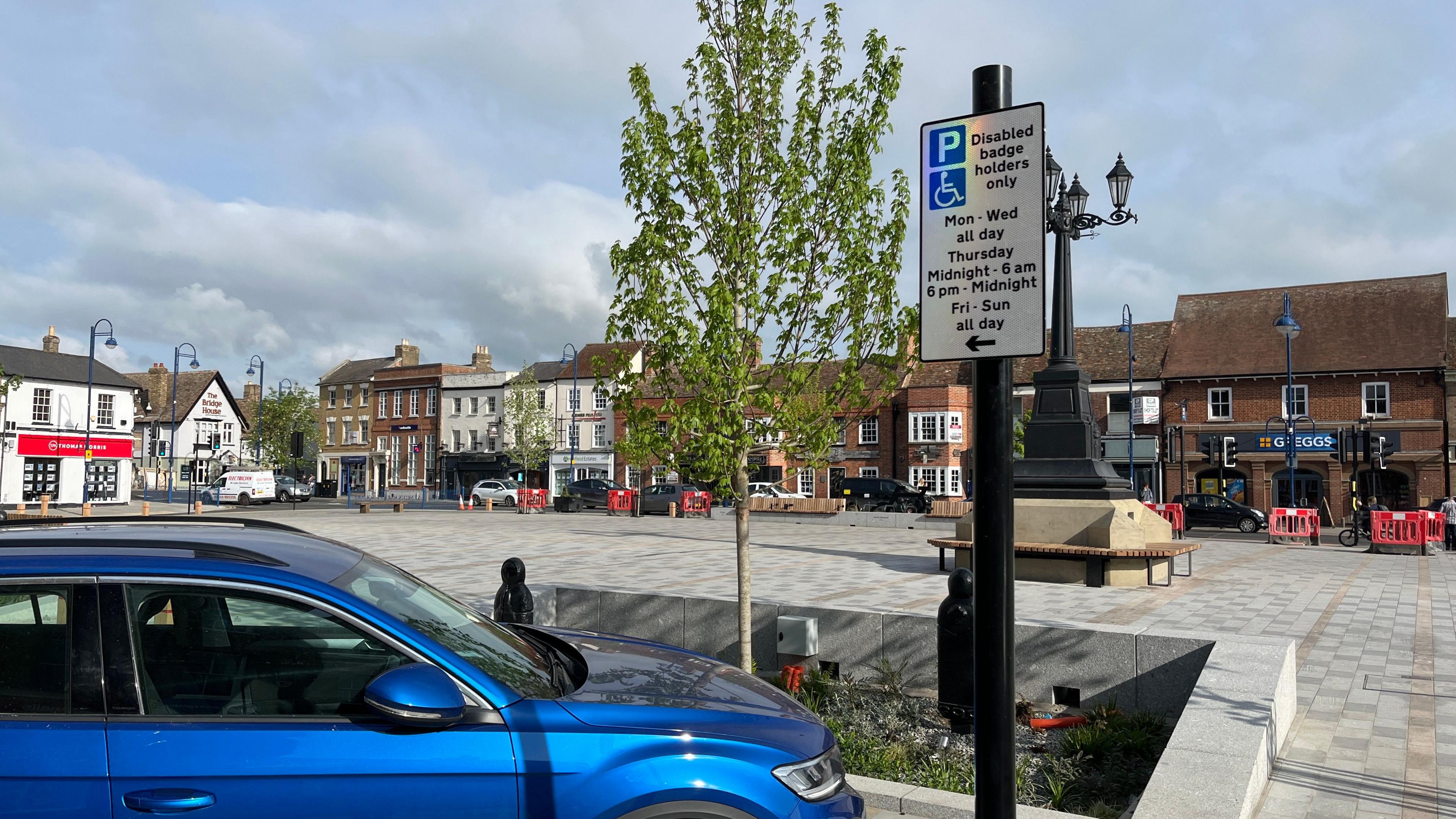 View of a blue car parked at the side of the market square. In front of it on a black pole is a sign stating "Disabled badge holders only".