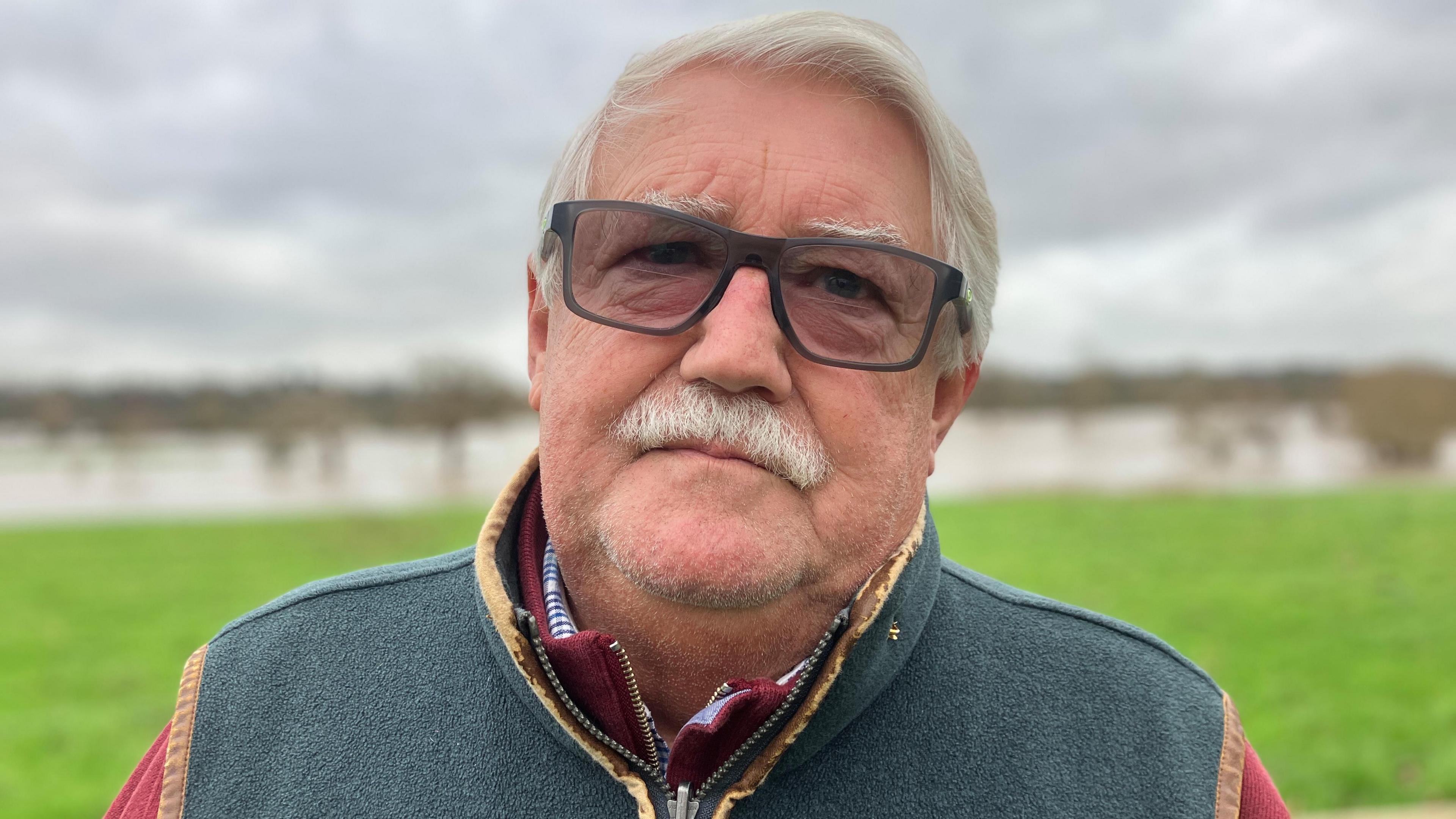 A man with glasses, white hair and a white moustache stands in front of partially-flooded field.