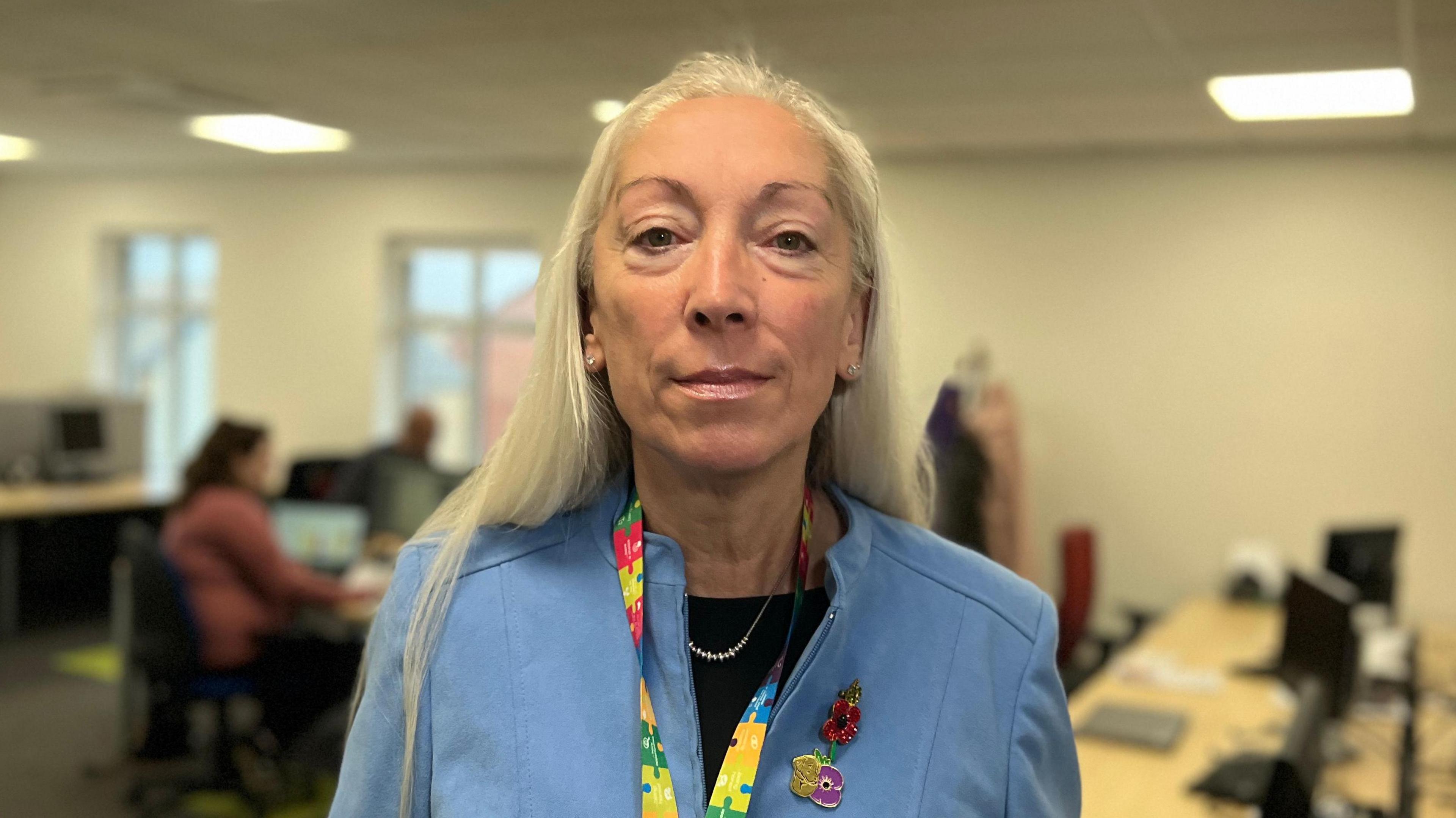 Sarah is wearing a pale blue jacket and a lanyard made up of colourful jigsaw shapes. On her lapel is a red metal poppy. Her hair is long and blonde and is worn down. She is looking directly at the camera. In the background, two people can be seen working at computers.