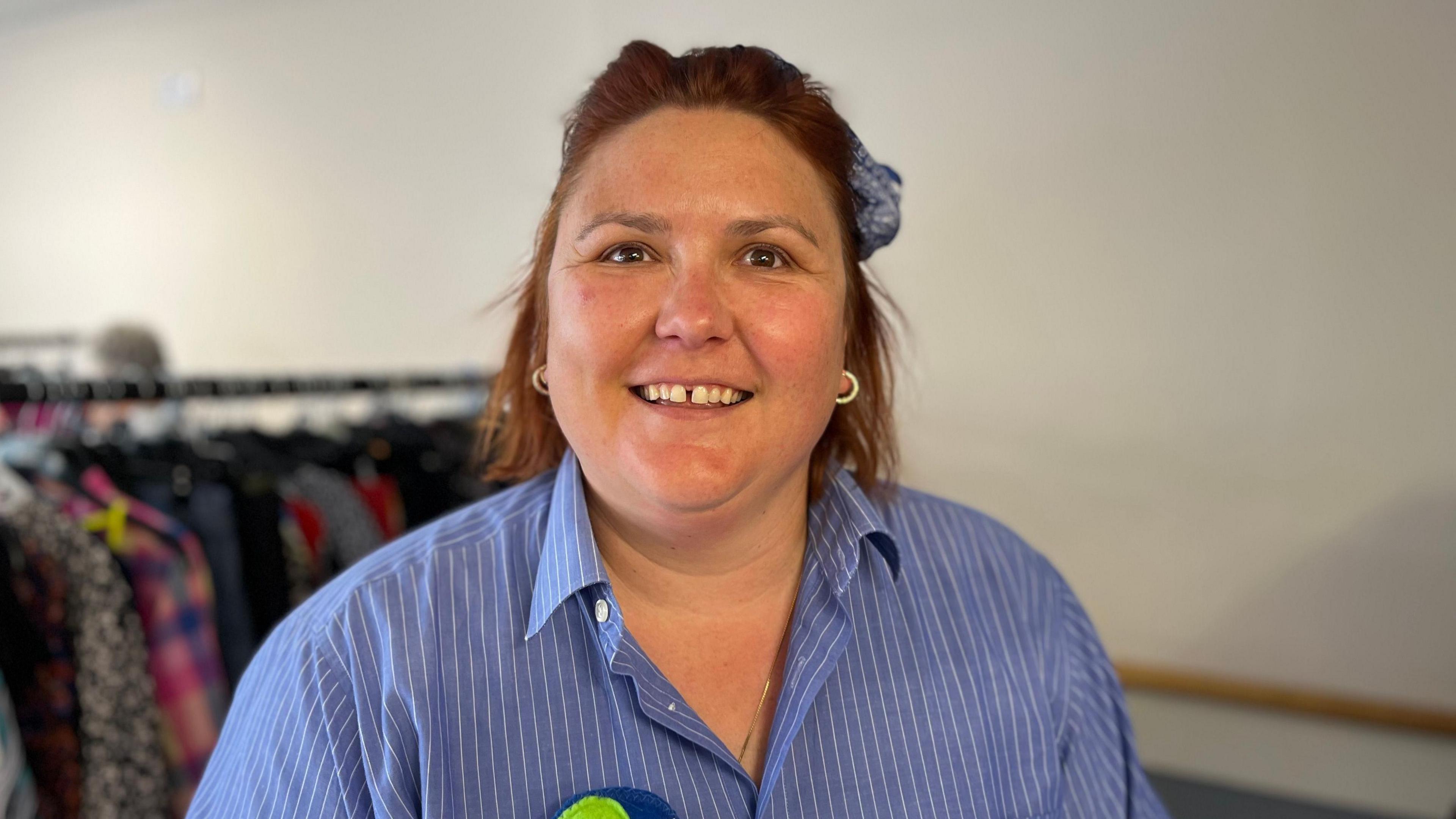 A lady with reddy brown hair looking towards the camera. She has a blue bow in her hair and is wearing earrings. She is wearing a blue and white striped shirt.
