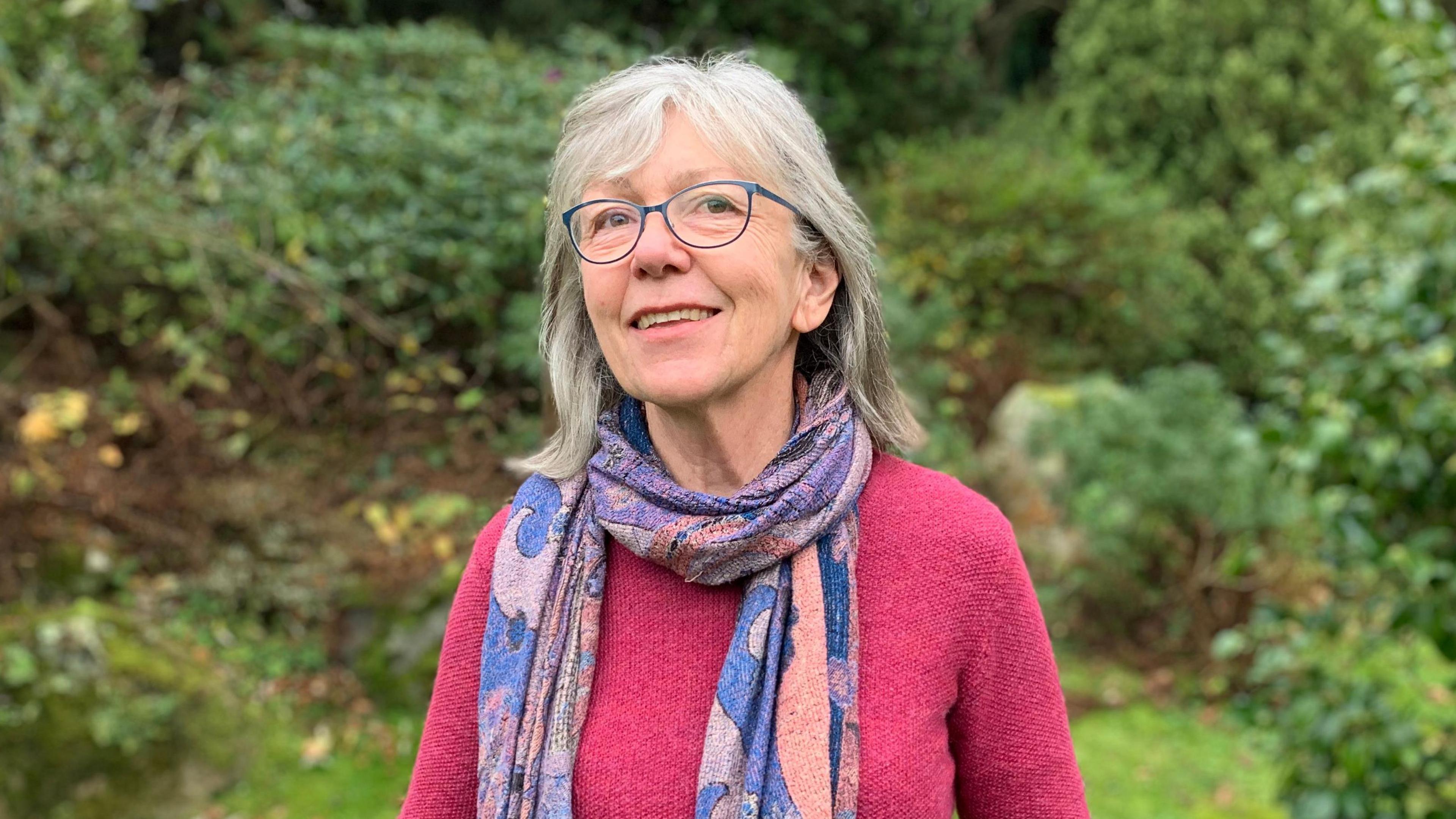 A woman with shoulder length grey hair wearing blue glasses a blue and purple scarf, on top of a pink jumper. 