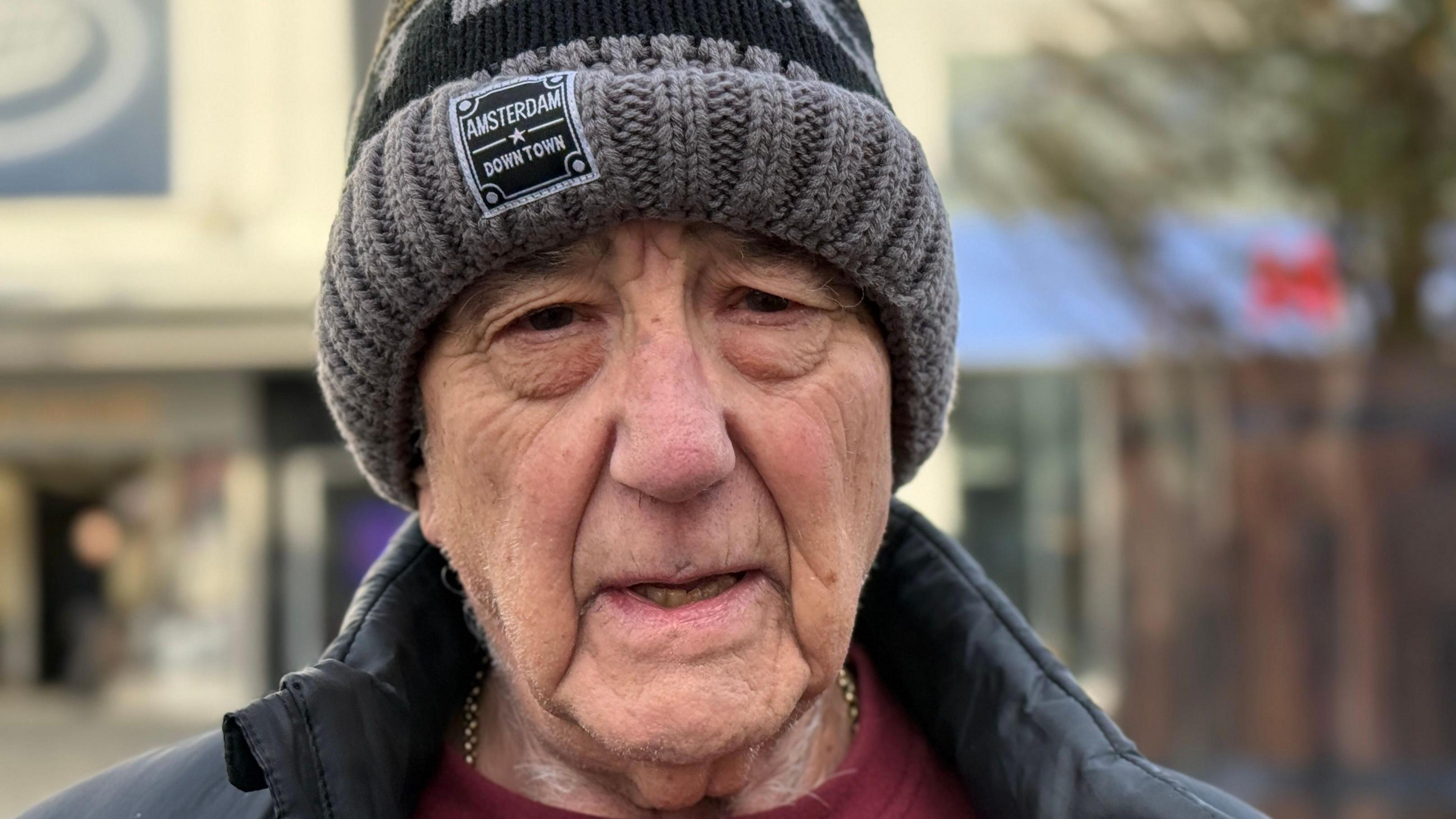 Garry Garland is looking at the camera and not smiling. He wears a black coat, red top and gold chain around his neck. He also has a grey and black woolly hat. He is stood in front of the controversial tree.