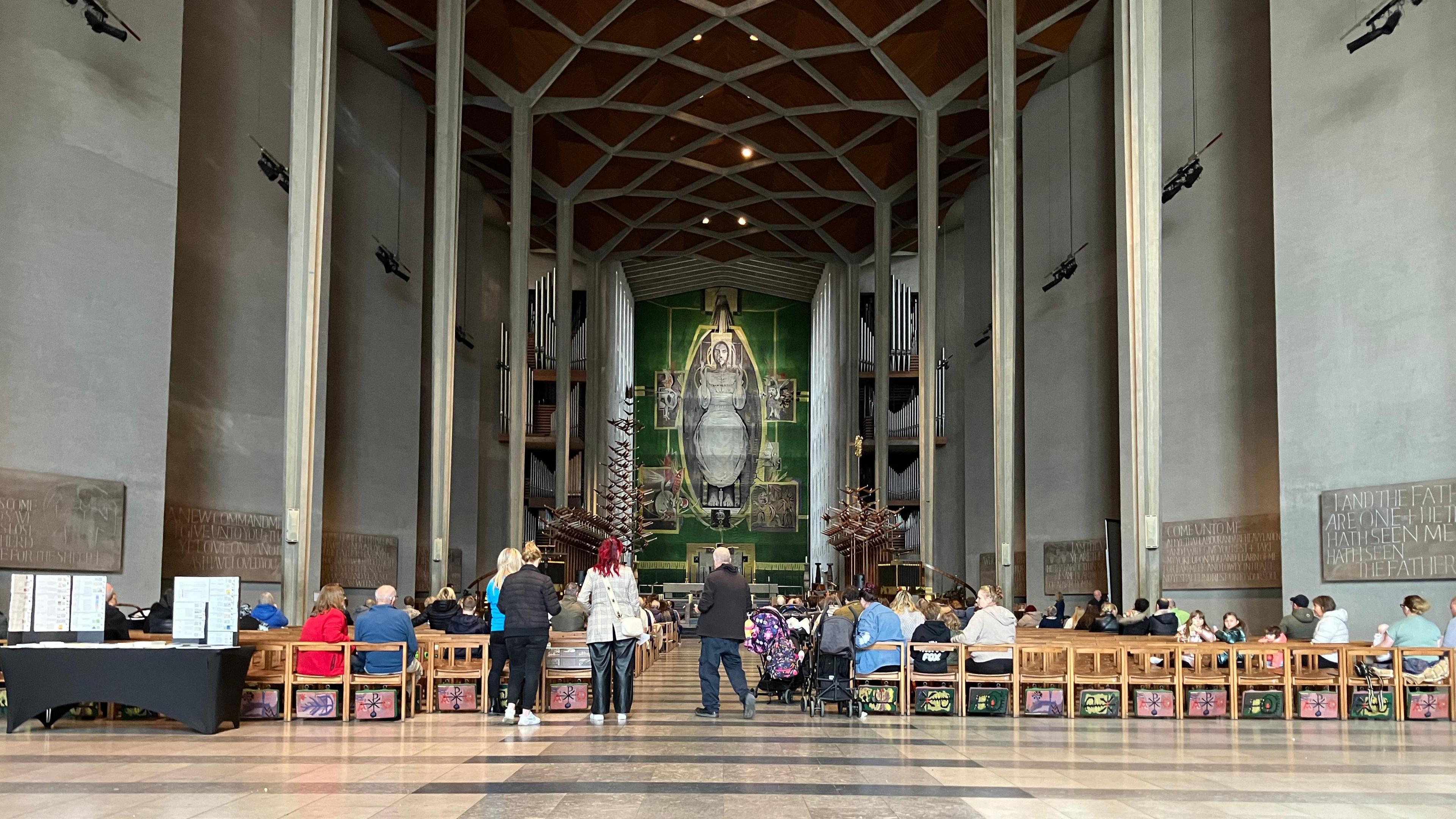 The interior of the cathedral, with tall, grey walls on either side and people standing or sitting in rows of chairs, leading down to the altar