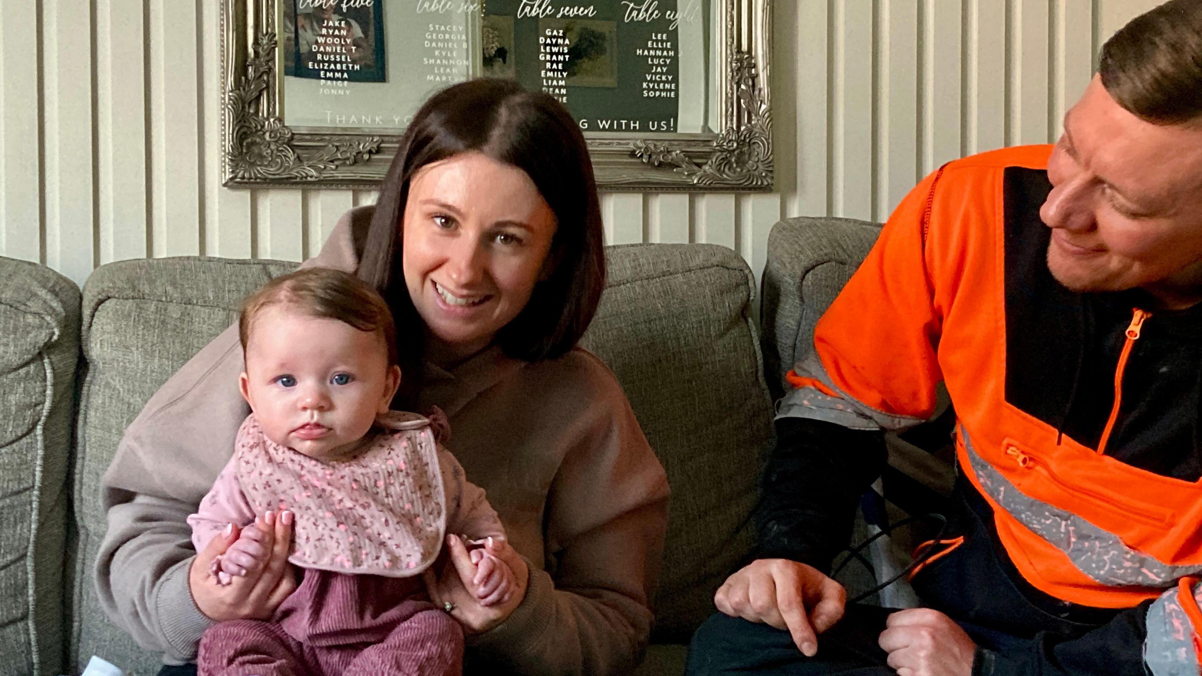 Emma Fellows, with a baby dressed in pink and wearing a bib on her knee, and her husband Jordan sitting next to her on the sofa. Ms Fellows is smiling at the camera and holding the girl's hands, and Mr Fellows is smiling at mother and daughter.