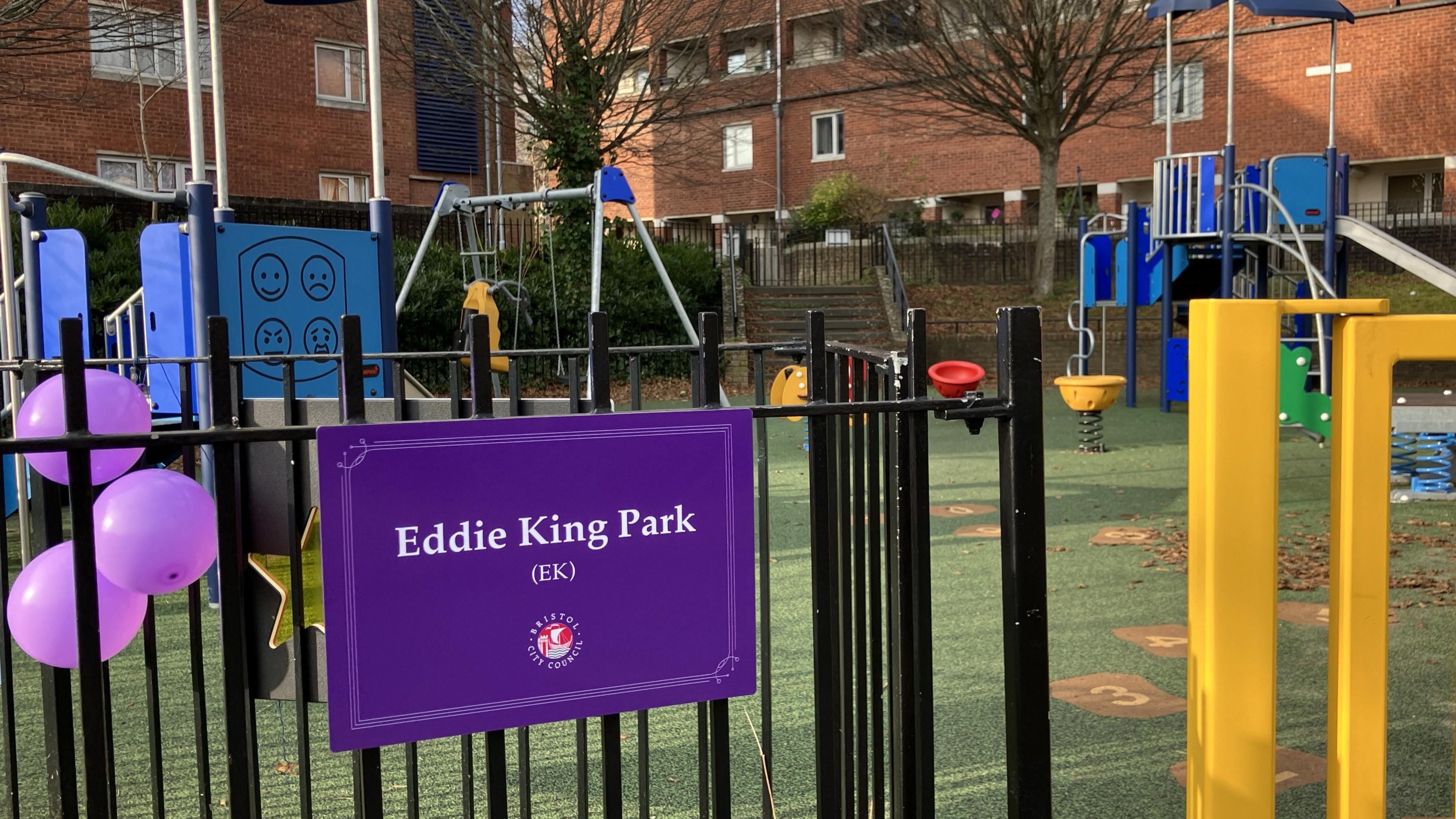 A close up shot of the black metal railings around the perimeter of the play park. Attached to the railings is a rectangular purple sign which says 'Eddie King Park' along with the Bristol City Council logo. Beside the sign there are three purple balloons tied to the railings. Inside the park you can see a swing set, climbing frames, see-saw and slide.