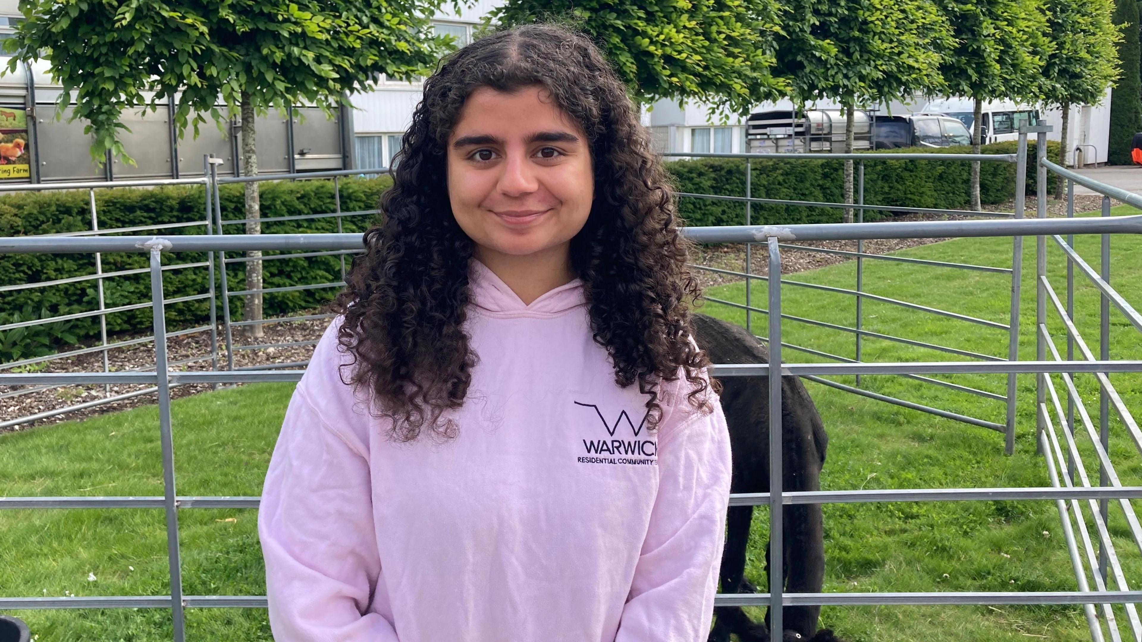 A young woman in a pink Warwick University hoodie and long curly black hair is standing in front of a pen where a black Shetland pony is eating the grass beneath. There is a row of trees in the background.