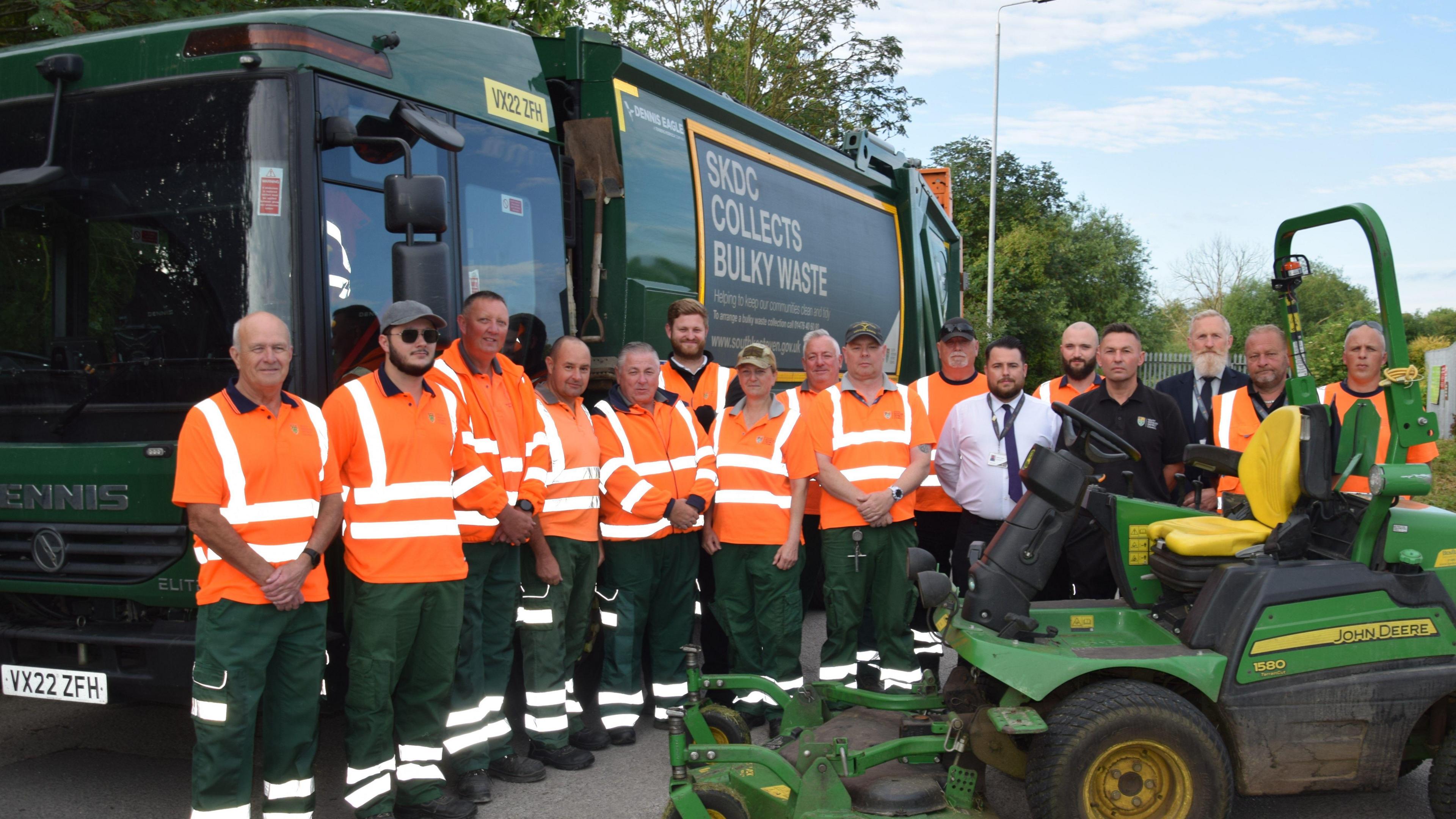 A group of uniformed workers in hi-vis orange tops, standing in front of a green bin lorry. There is a lawnmower in the foreground. There are trees in the background. The sky is blue with light clouds.