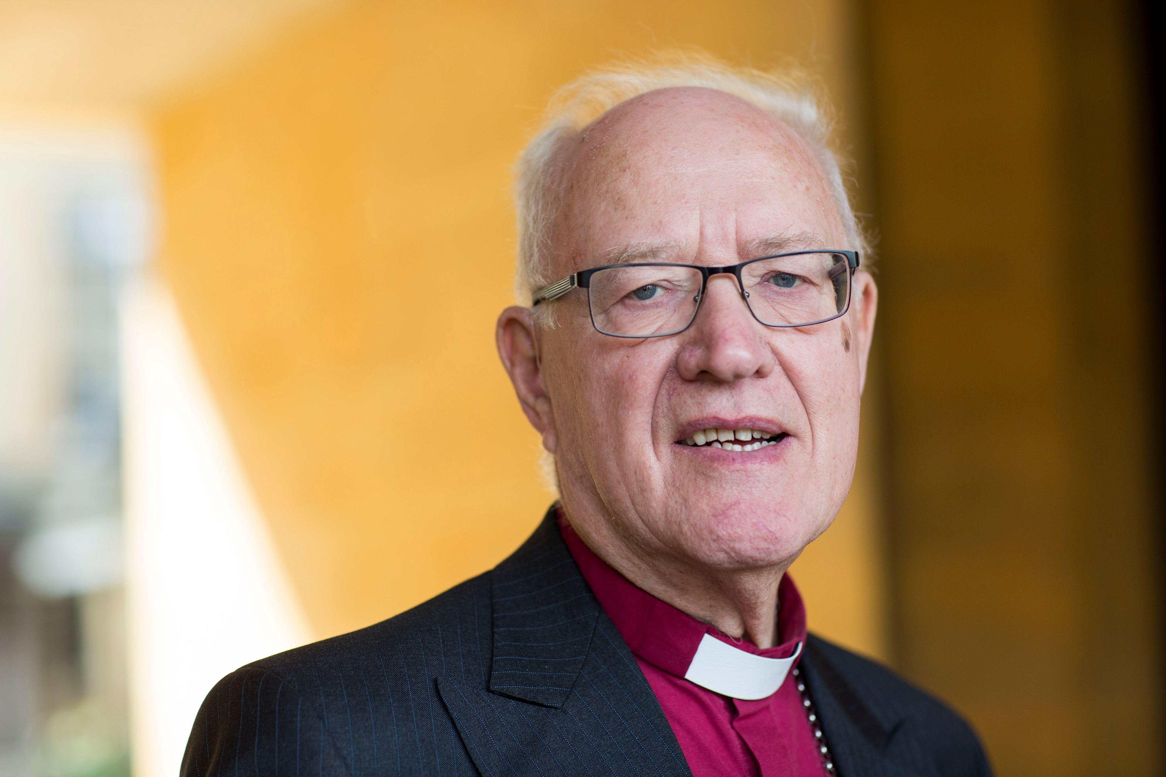 Portrait of George Carey, who has balding white hair and black-framed glasses. He is wearing a purple clerical shirt with white clerical collar and a black pinstripe jacket. He is pictured outside in front of a yellow-painted wall. 