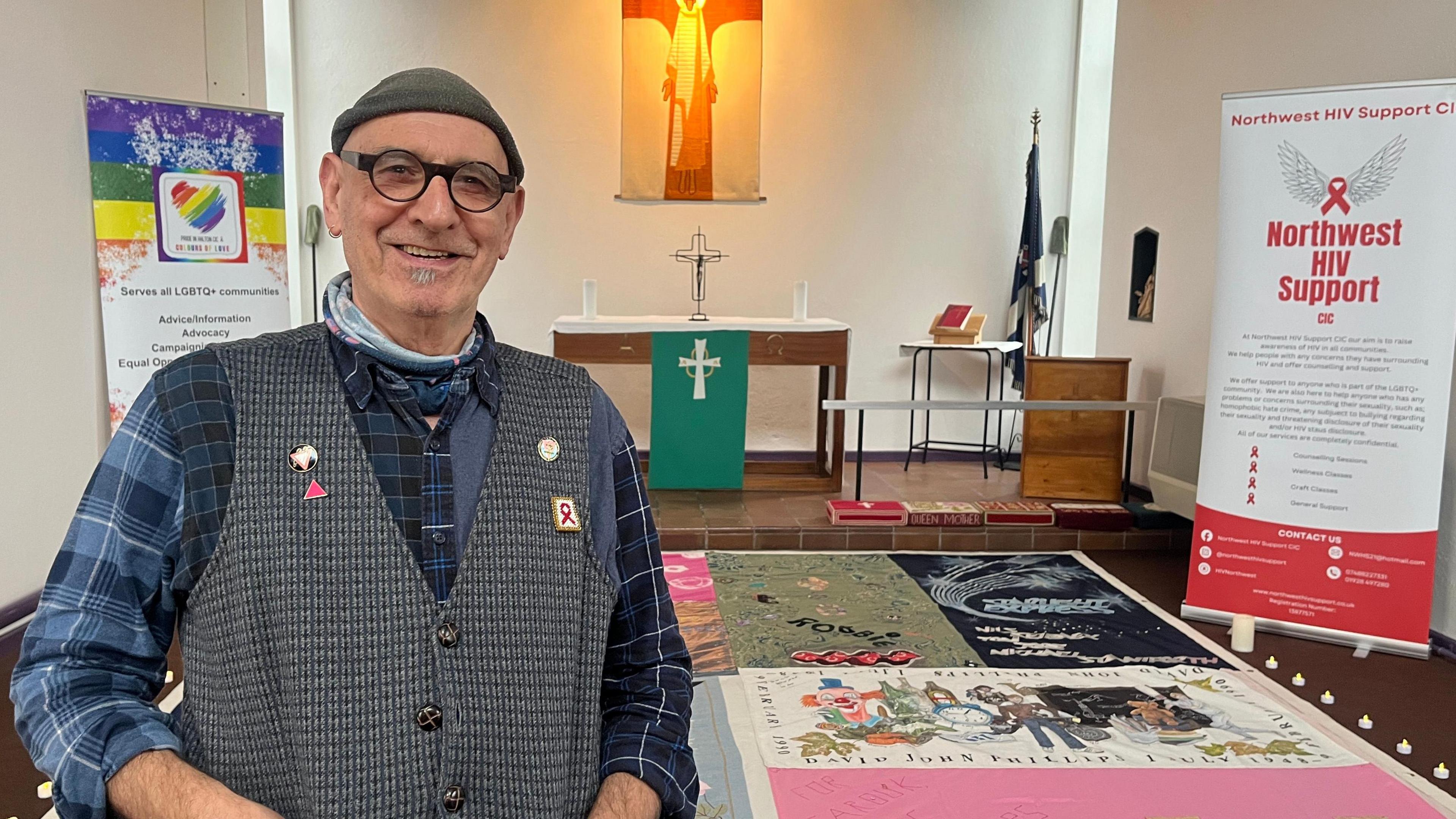 Jonathan Blake wearing a black beanie hat blue black and white checked shirt and grey tweed waistcoat with badges one with the HIV/Aids red ribbon smiling inside St Andrew's Church with the memorial quilt panel laid on the floor