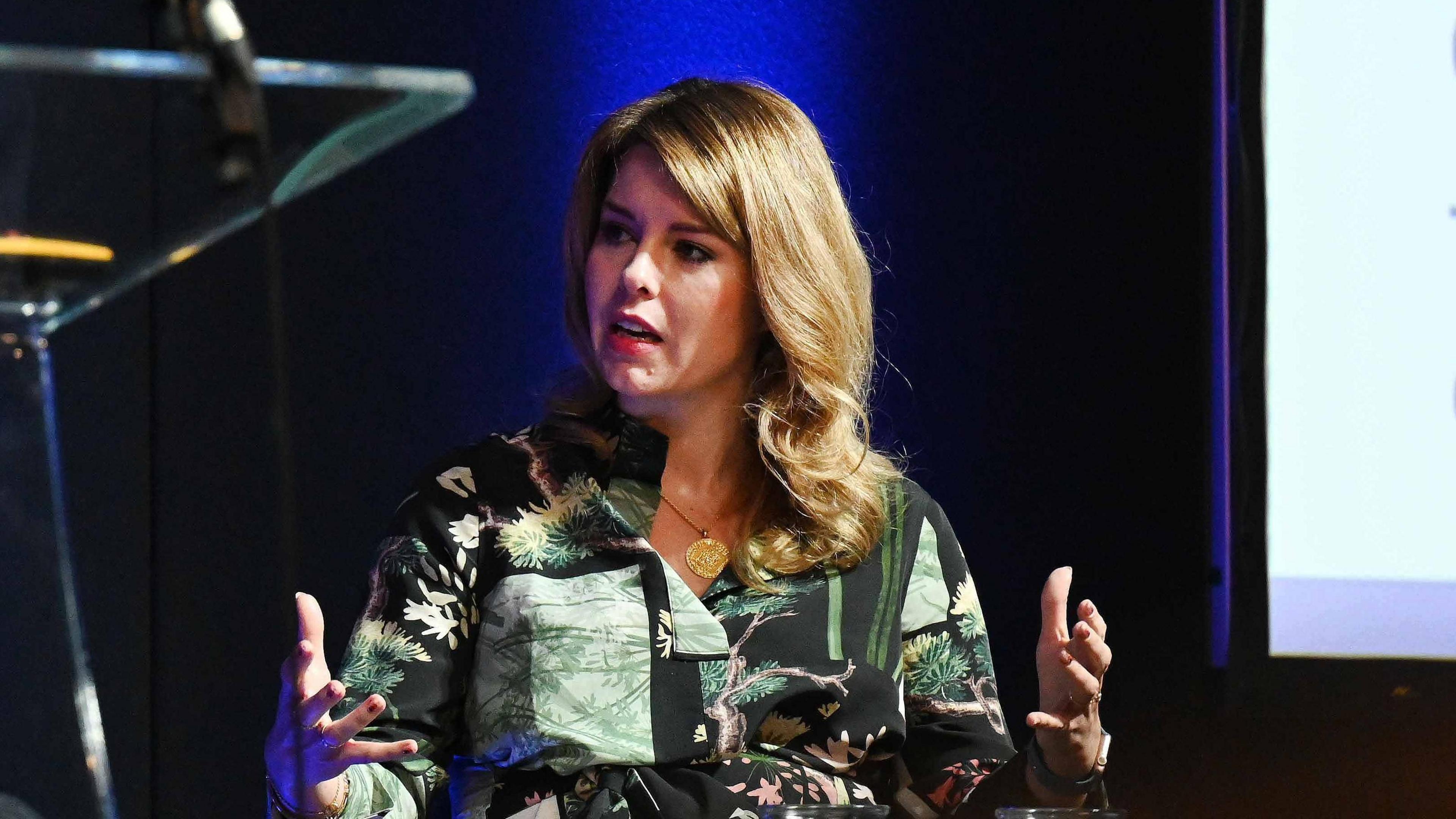 Kim McGuinness sitting on a chair with her hands in the air. She is wearing a patterned green top of tree images and has blonde hair. 