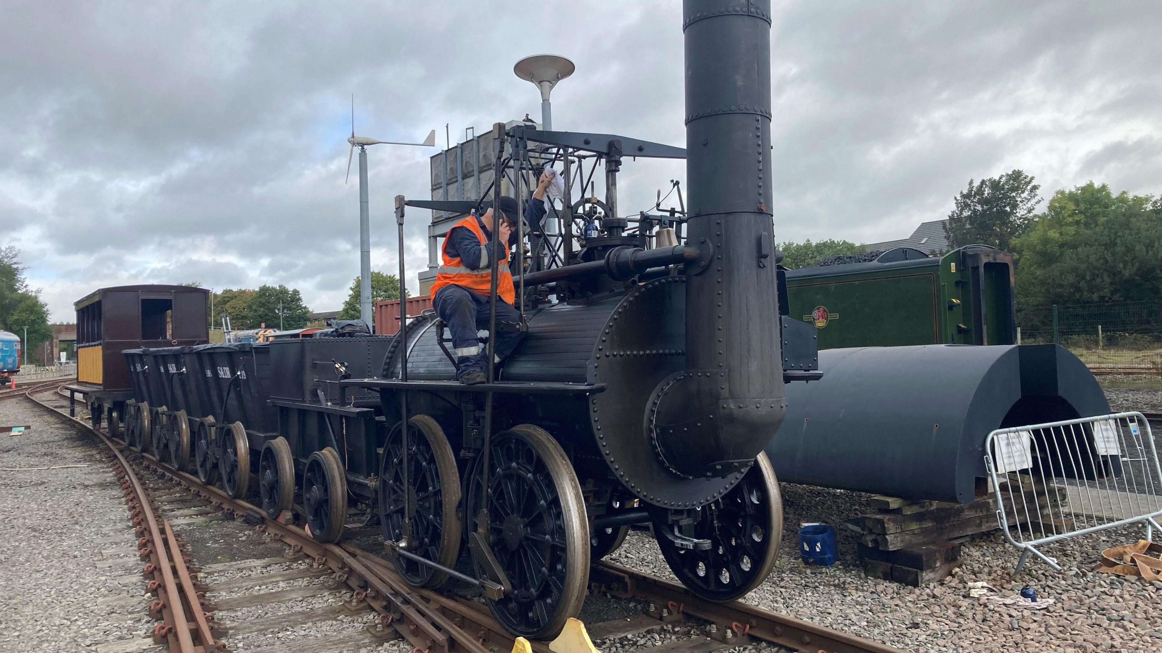 A man in orange high-vis sits on a black steam engine consisting of a large boiler, funnel, pistons and four large wheels on a section of track. Behind the engine are four black coal wagons and a covered passenger cart.