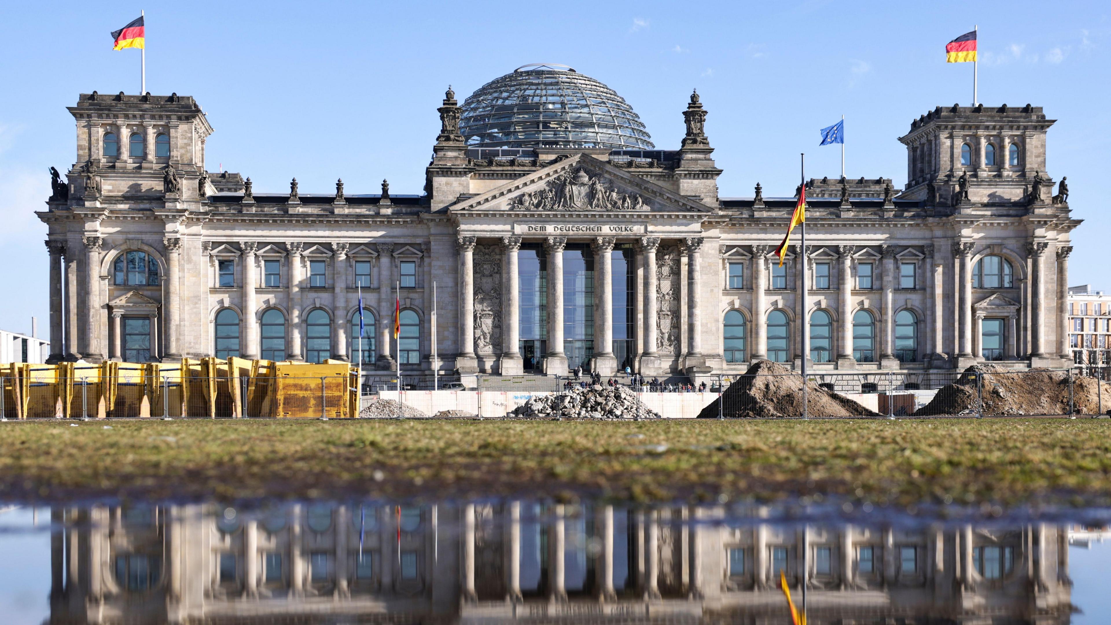 Reichstag building in Berlin, a large of white building with glass windows and doors. It has a glass dome on top with two German flags flying, one from each side. There is a further German flag displayed in front of the building. A European union flag is also on the building. There is work in progress in front of the building. The sky is blue. 