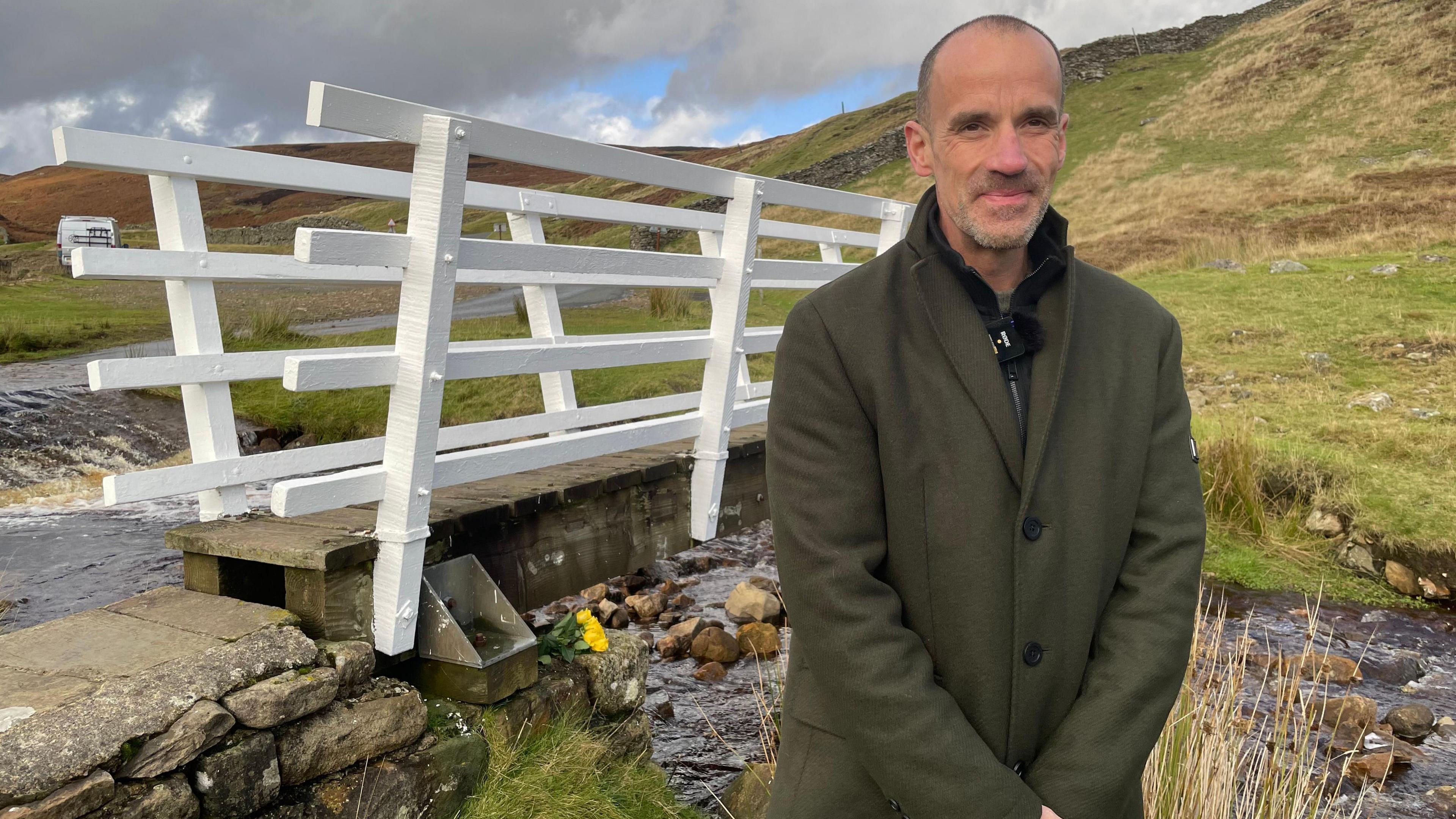 Peter Lefley standing by the bridge in the Yorkshire Dales, wearing a green wool coat 