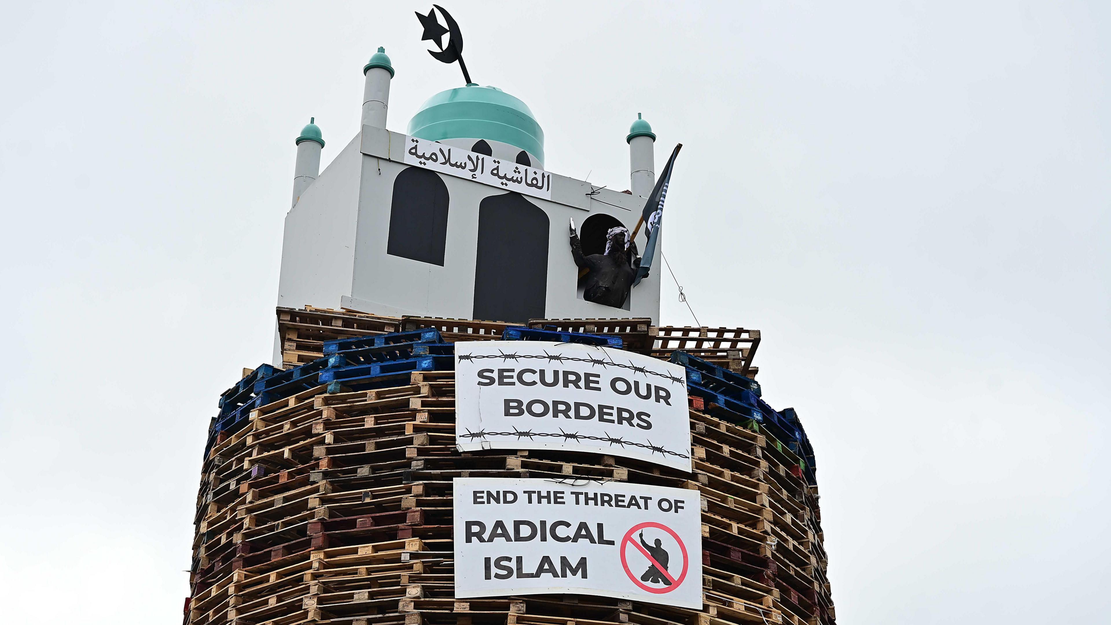 An image of the top of the bonfire, consisting of wood pallets, with a structure resembling a mosque on top. Two placards are below. One reads "secure our borders" while another reads "end the threat of radical Islam". 
A figure of a man is coming out from one of the windows. The figure is wearing a headscarf and is holding a model of a knife and a flag.