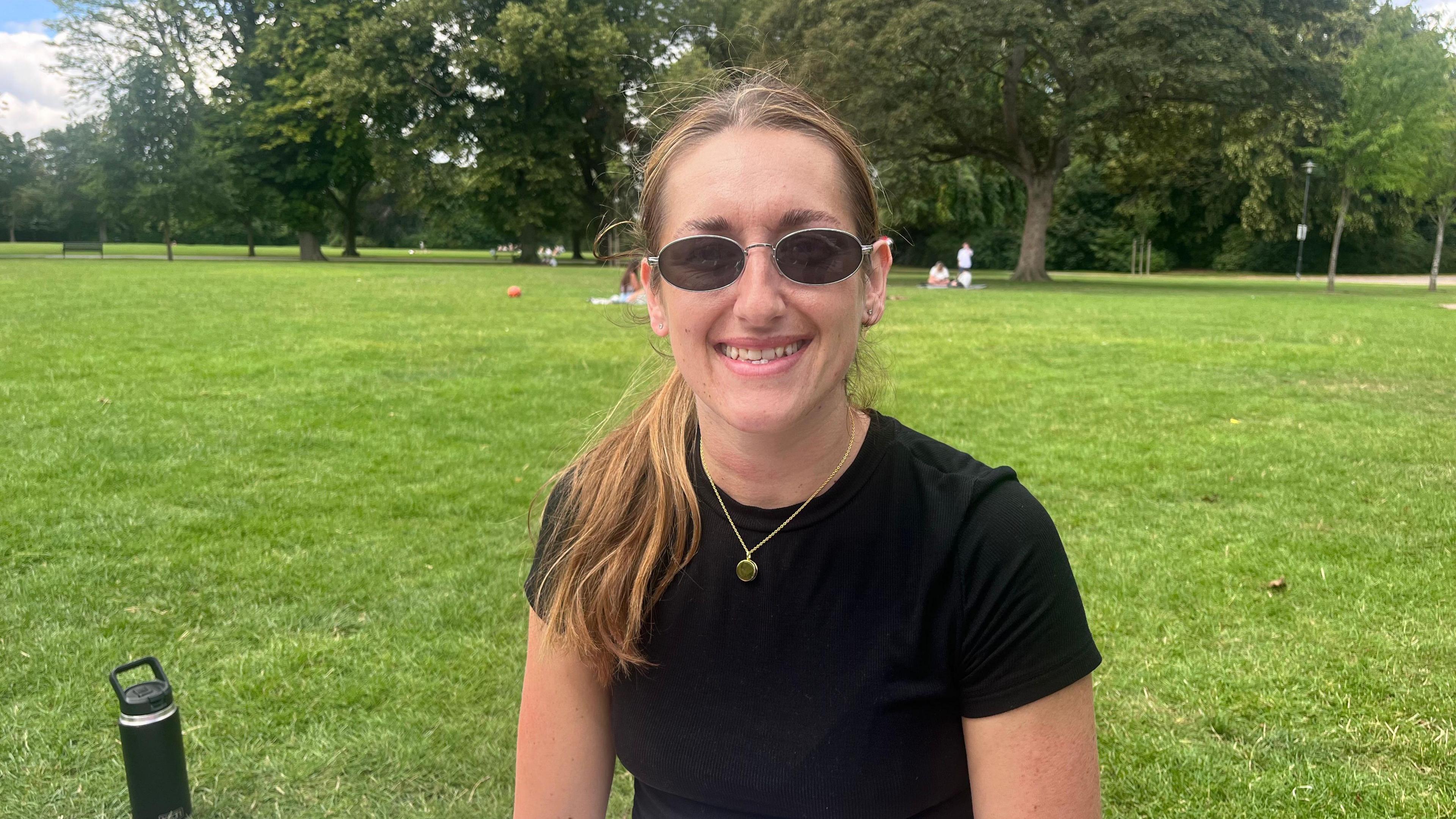 Daisy Robson sits in a green field staring at the camera with sunglasses on and wearing a black T-shirt.