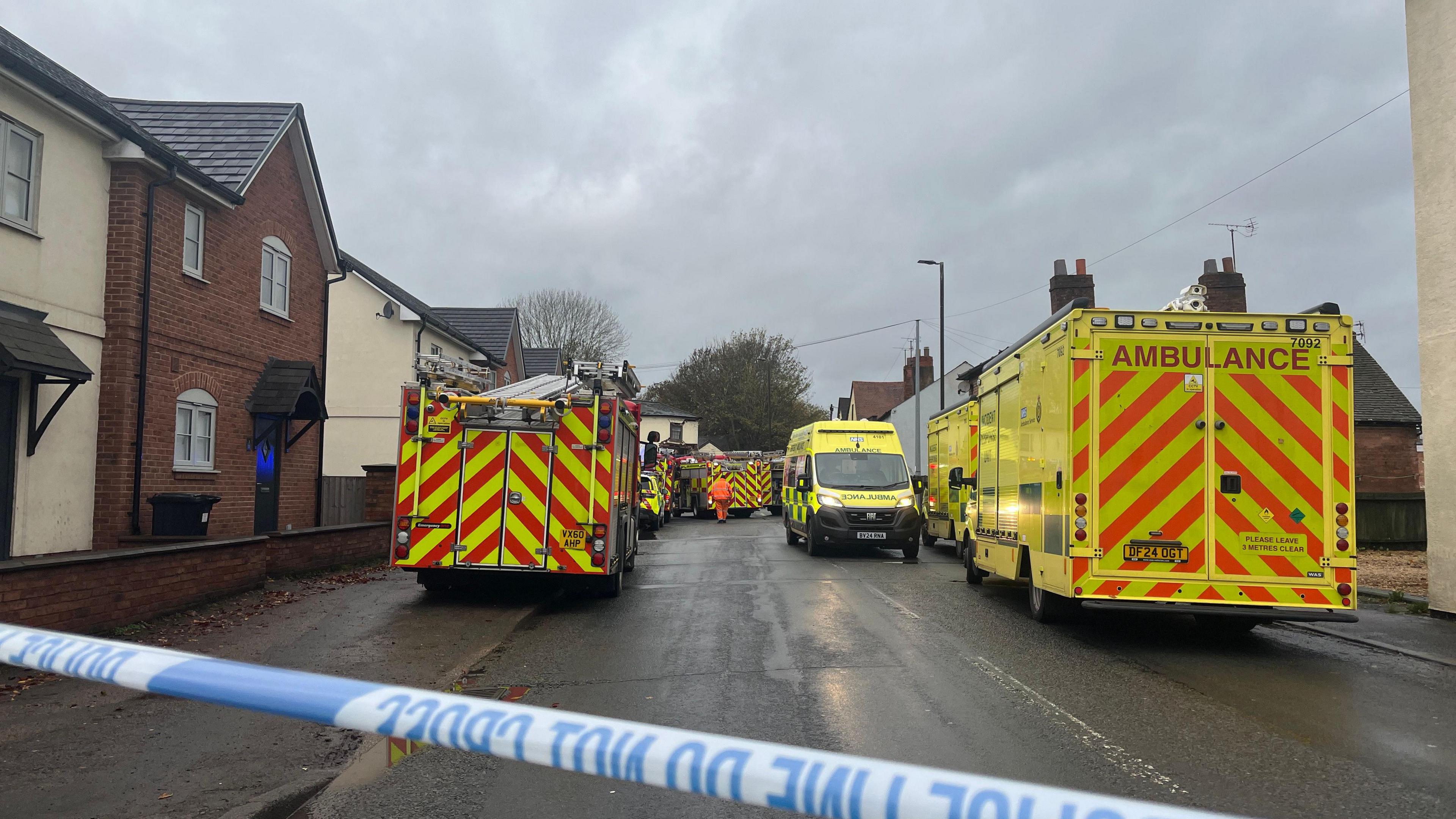A fire engine and three ambulances are parked on a residential road while police tape prevents people from entering the scene. 