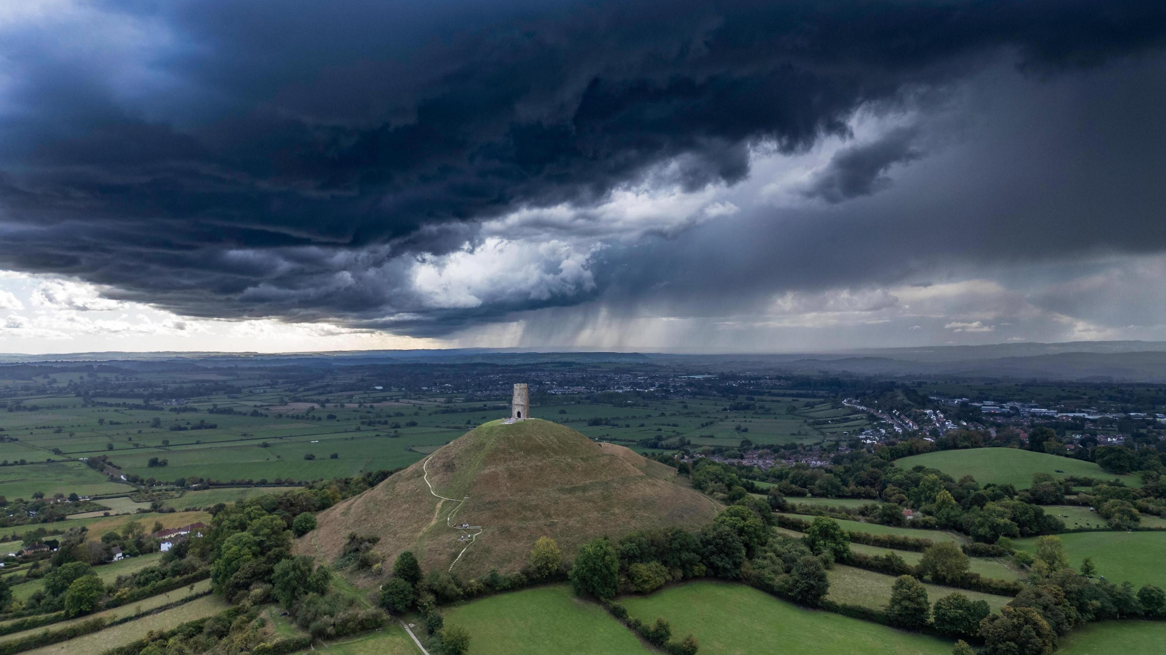 Large black clouds gather above Glastonbury Tor, which is a prominent hill overlooking green fields that stretch as far as you can you. The roofs of houses can be seen in the distance. 