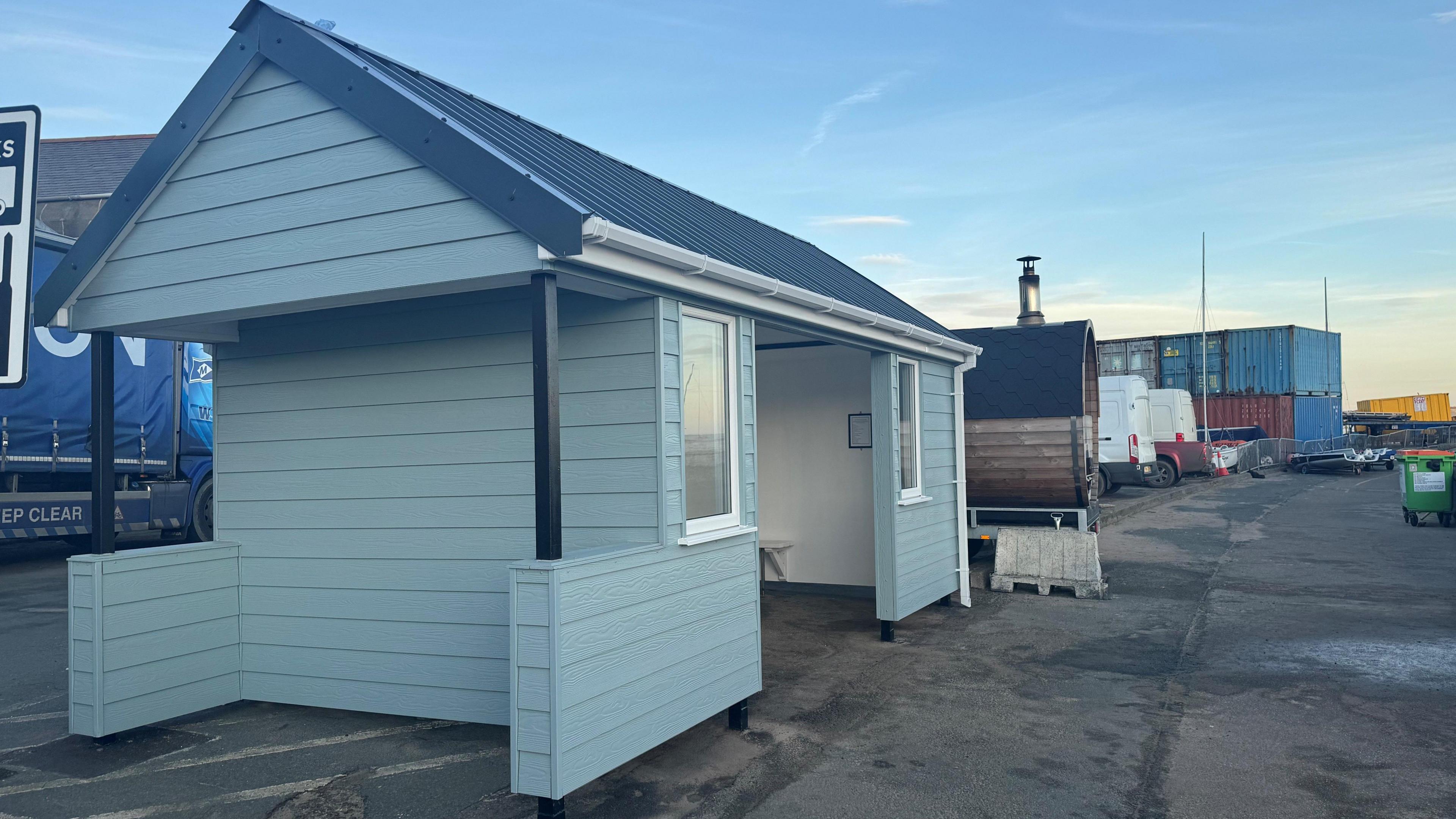 The hut is duck egg blue, it has two small windows and a wide entrance. It has a grey roof. It stands on grey tarmac next to a beach sauna.