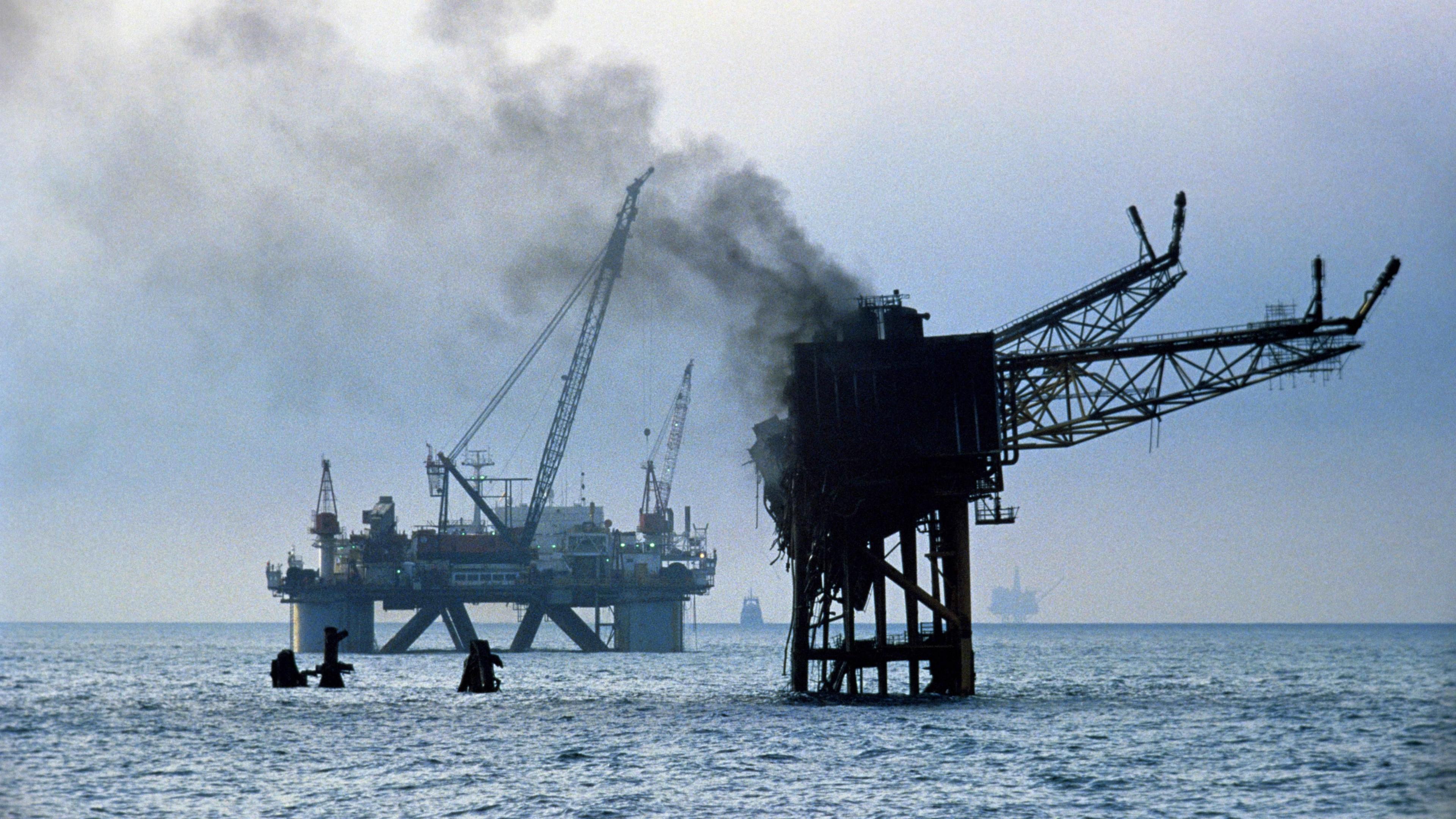 The smoking remains of the Piper Alpha oil and gas production platform. The structure is blackened and buckled in silhouette, standing in calm seas. Two platforms and a boat can be seen in the background. 