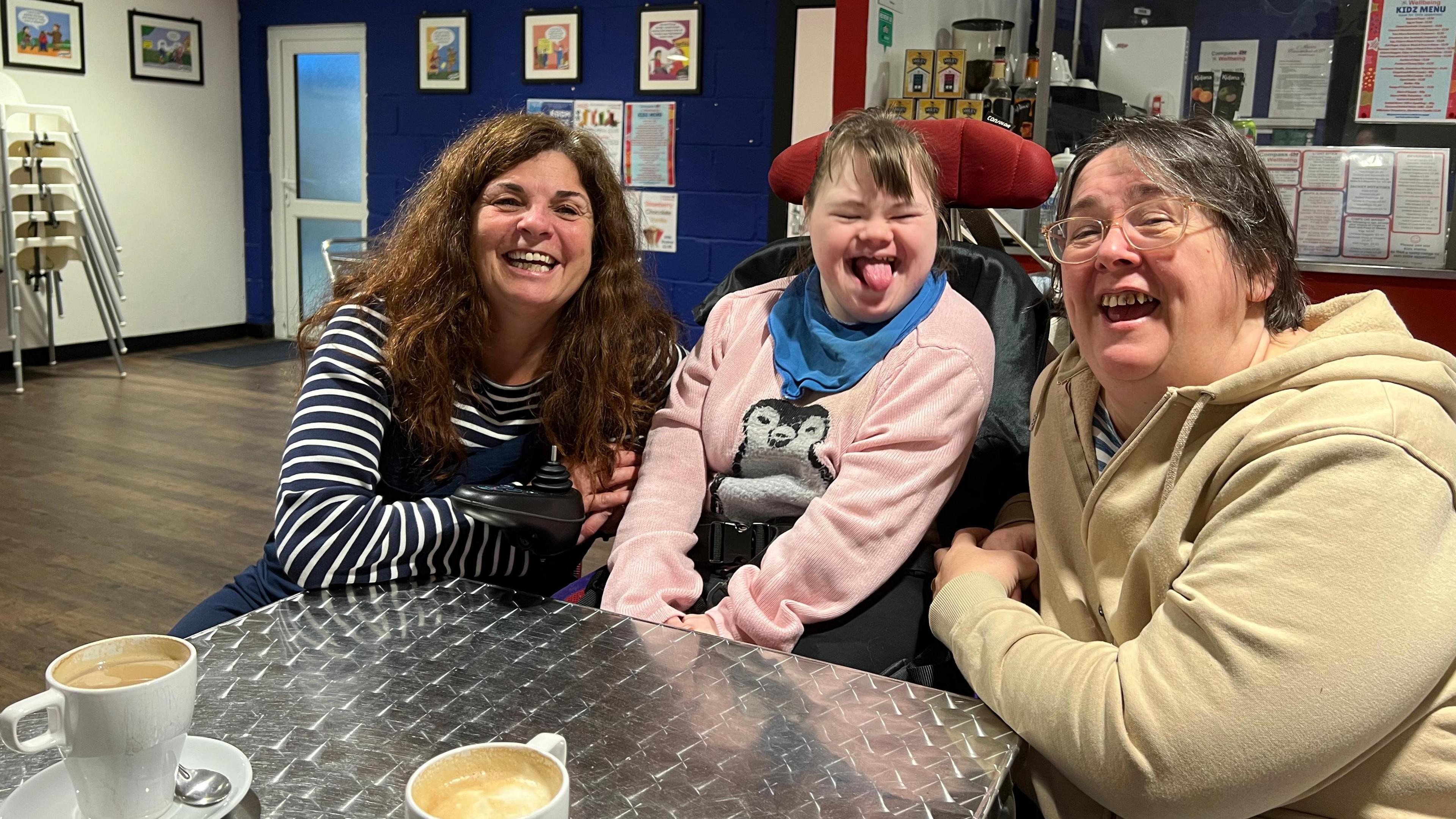 Three women are sitting at a metal table and laughing at the camera. There are coffees on the table.