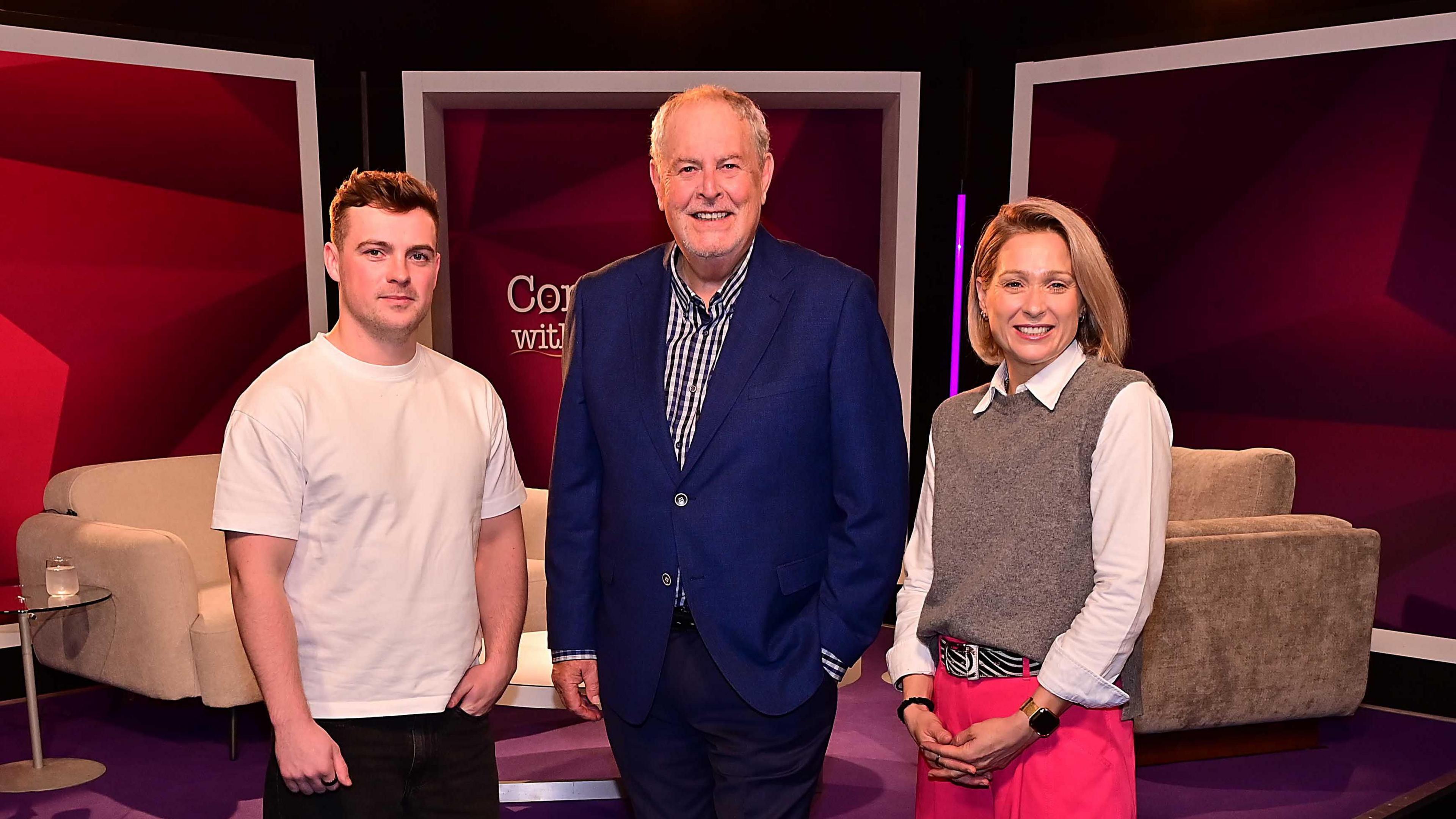 Three people standing beside each other, two men and a woman. The man has short dark hair and is wearing a white t-shirt. The man has short grey hair and is wearing a blue blazer and checked shirt. The woman has shoulder length blonde hair and is wearing pink trouser, white shirt and brown jumper.