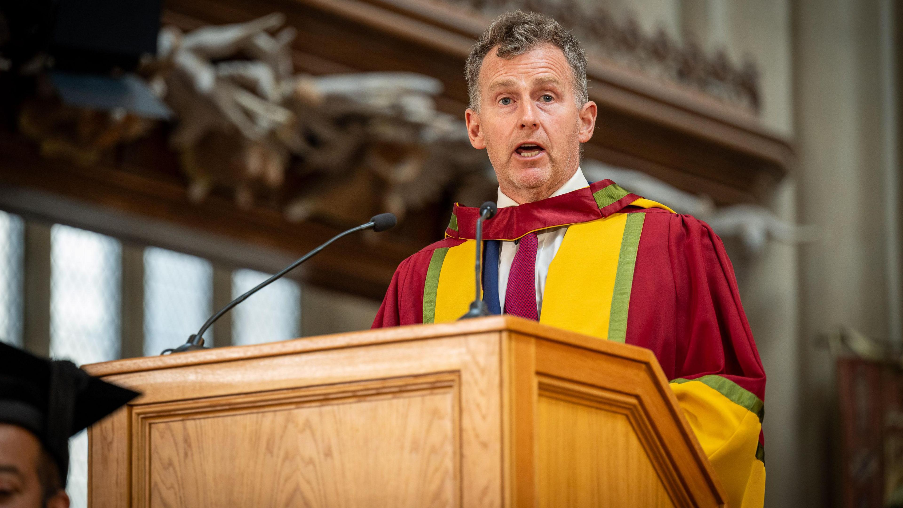 A man wearing red graduation robes standing behind a lectern