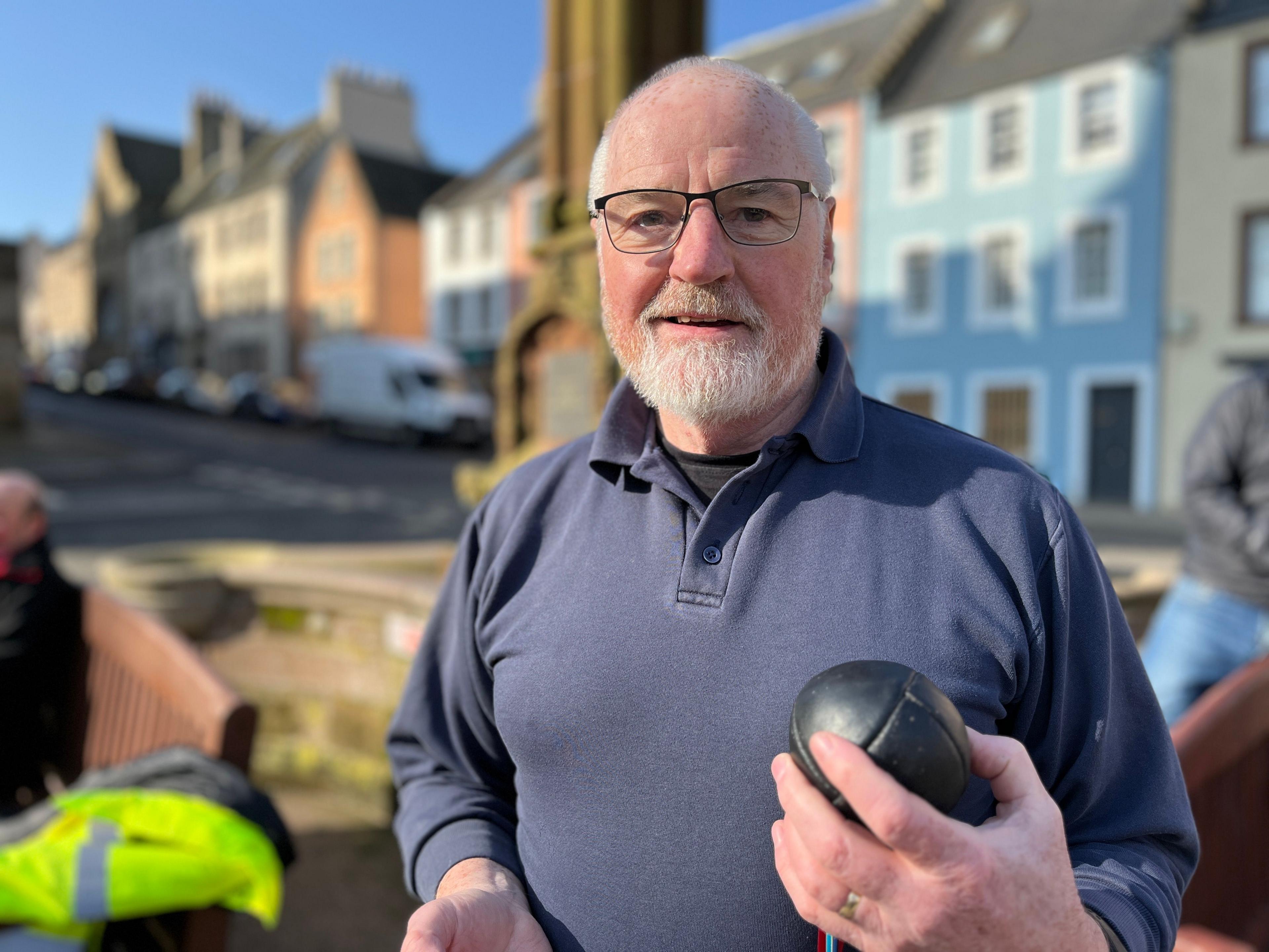 Former provost Len Wyse who is grey and balding with a beard and glasses. He is wearing a blue button-necked jumper and is carrying a small leather ball