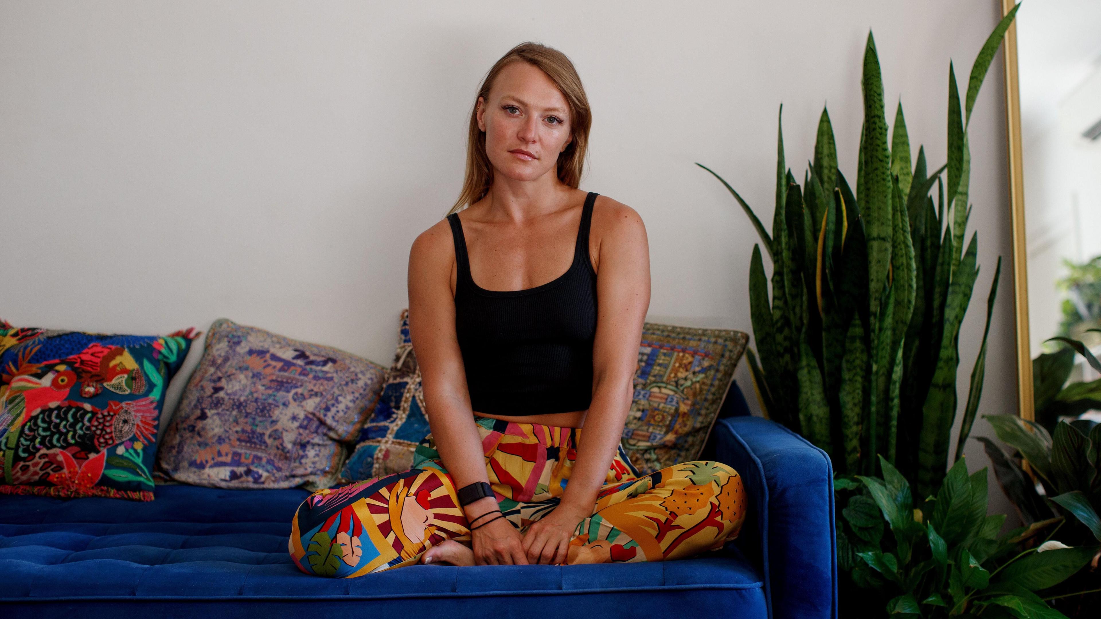 A woman in brightly patterned trousers sits on a blue sofa and stares at the camera. She isn't smiling. She is surrounded by patterned, colourful cushions and next to her are several plants