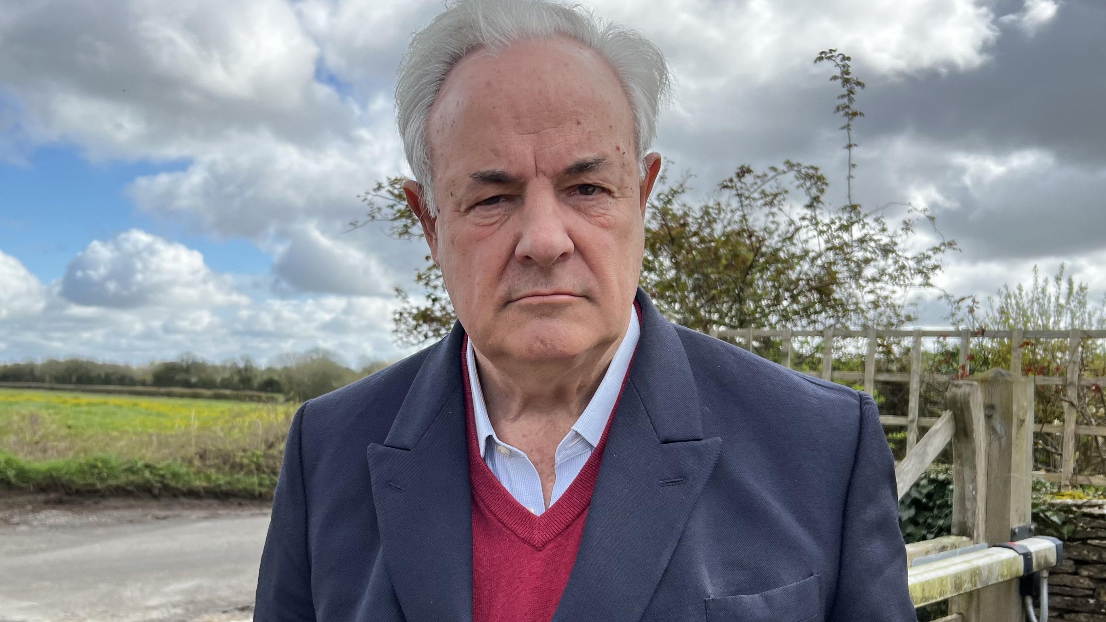 James Gray MP for North Wiltshire standing in front of a field gate wearing a blazer and shirt