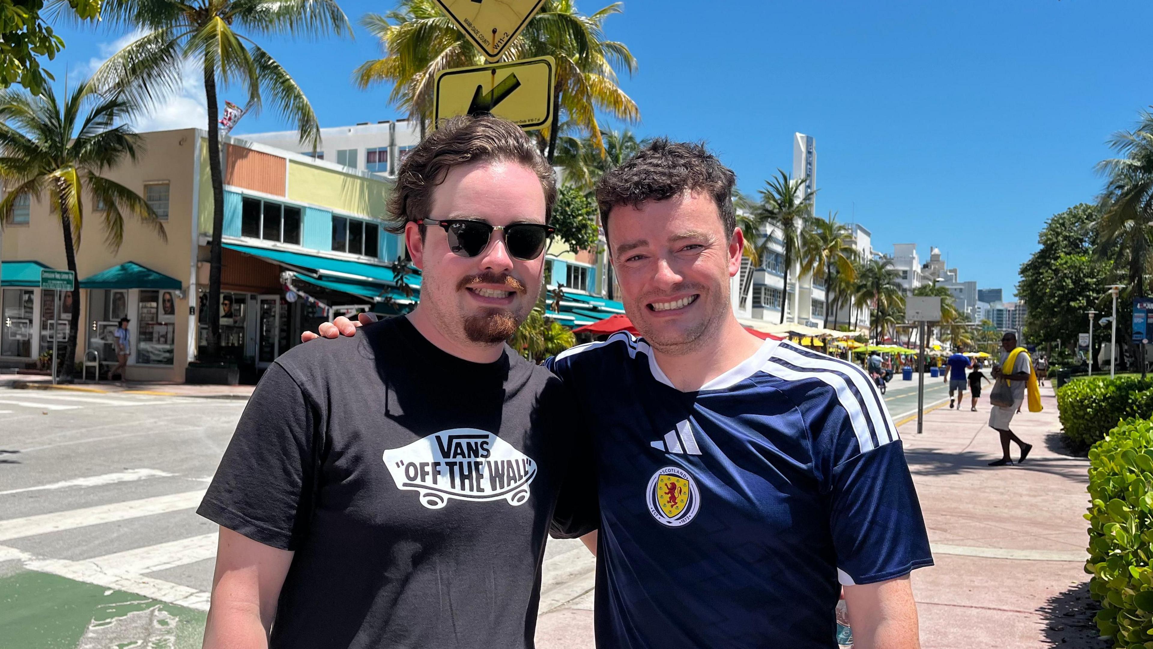 Two men with dark hair, one wearing a Scotland football top standing in a Miami street.